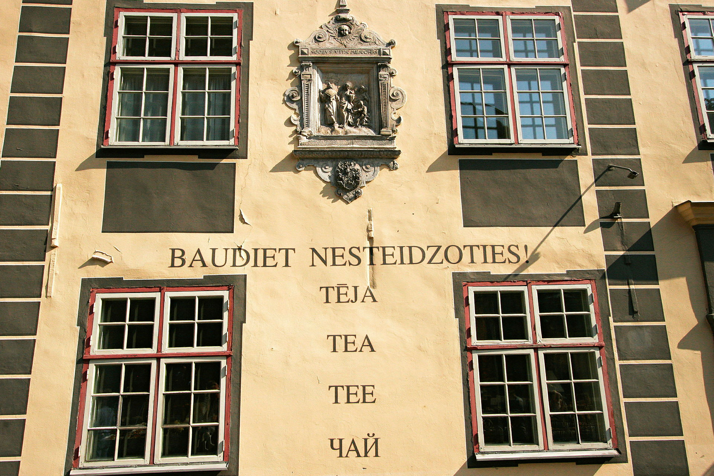 Fasade av en bygning med fire store vinduer, en dekorativ steinrelieff og teksten "Baudiet nesteidzoties! Tēja, Tea, Tee, Чай". (AI-generert tekst)