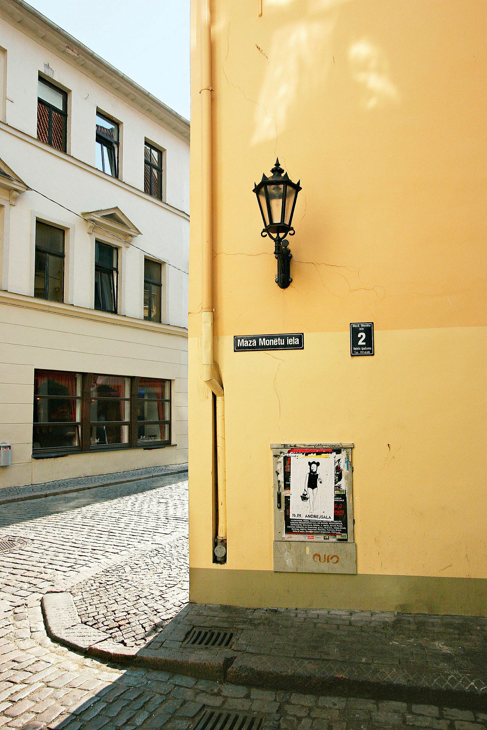 Et hjørne av en bygning med gul fasade, en gatelykt, et skilt som angir gaten 'Maza Monētu iela', og et område med plakater. (AI-generert tekst)