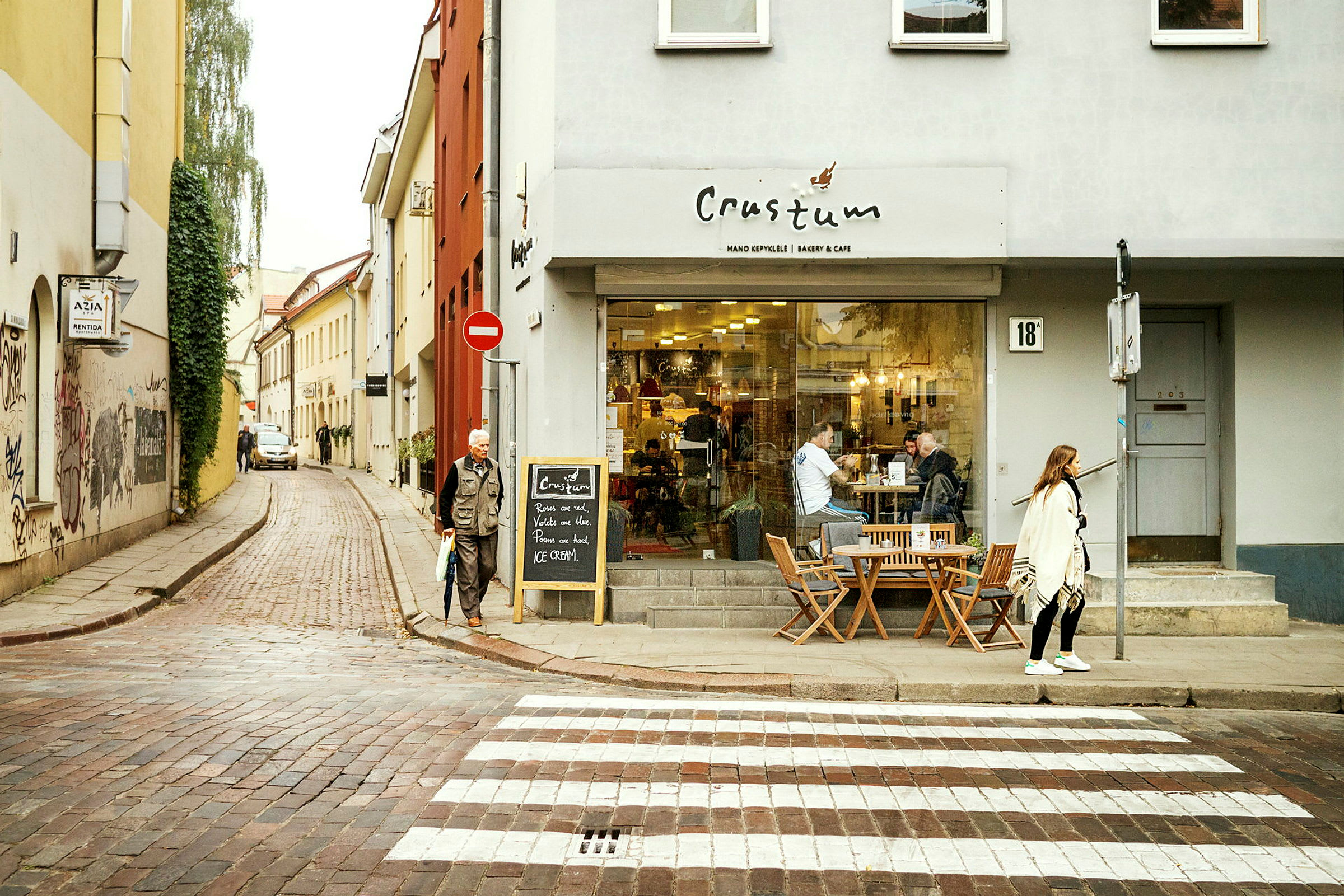 En gate med en kafé på høyre side. Mennesker sitter utendørs på kaféens uteservering, og en person krysser et fotgjengerfelt. (AI-generert tekst)