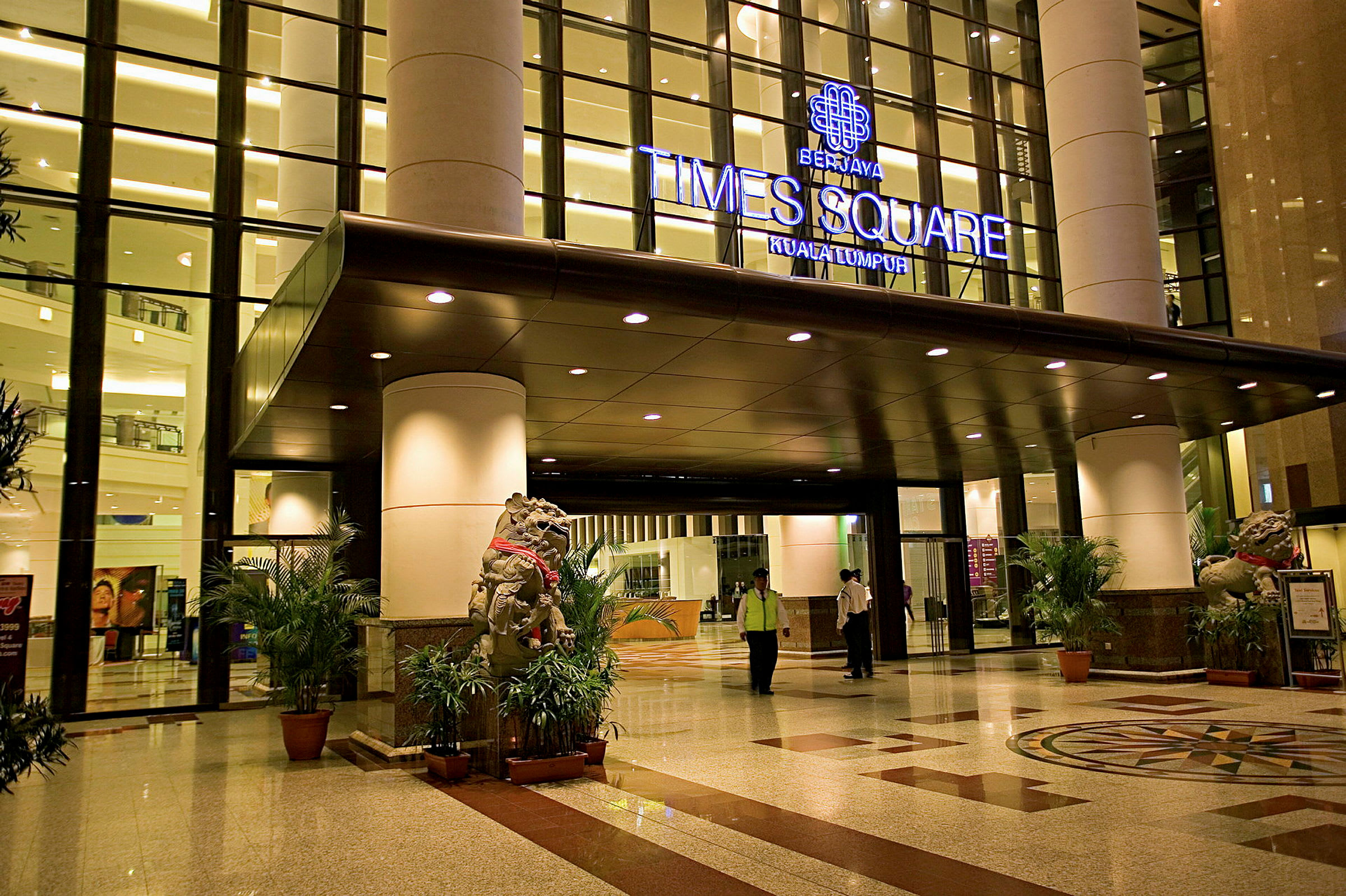 Inngangsområde til en bygning med glassfasade og lys belysning. Et skilt over inngangen angir 'Berjaya Times Square Kuala Lumpur'. Tregulv og dekorative planter er også synlige. (AI-generert tekst)
