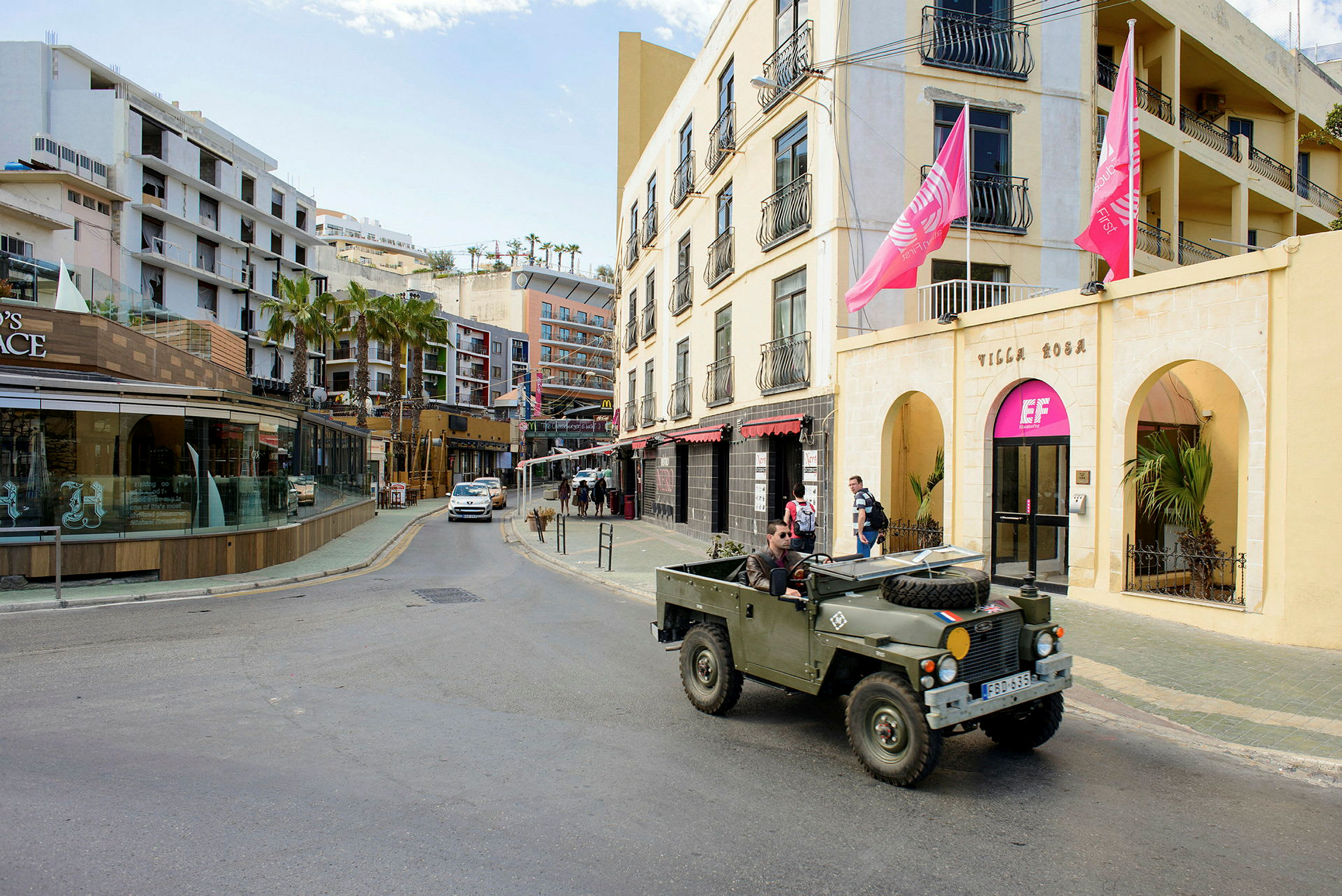 En gate med bygninger, inkludert en gul fasade med rosa flagg og en butikk med glassvinduer. En militærgrønn jeep er parkert ved veikanten. (AI-generert tekst)