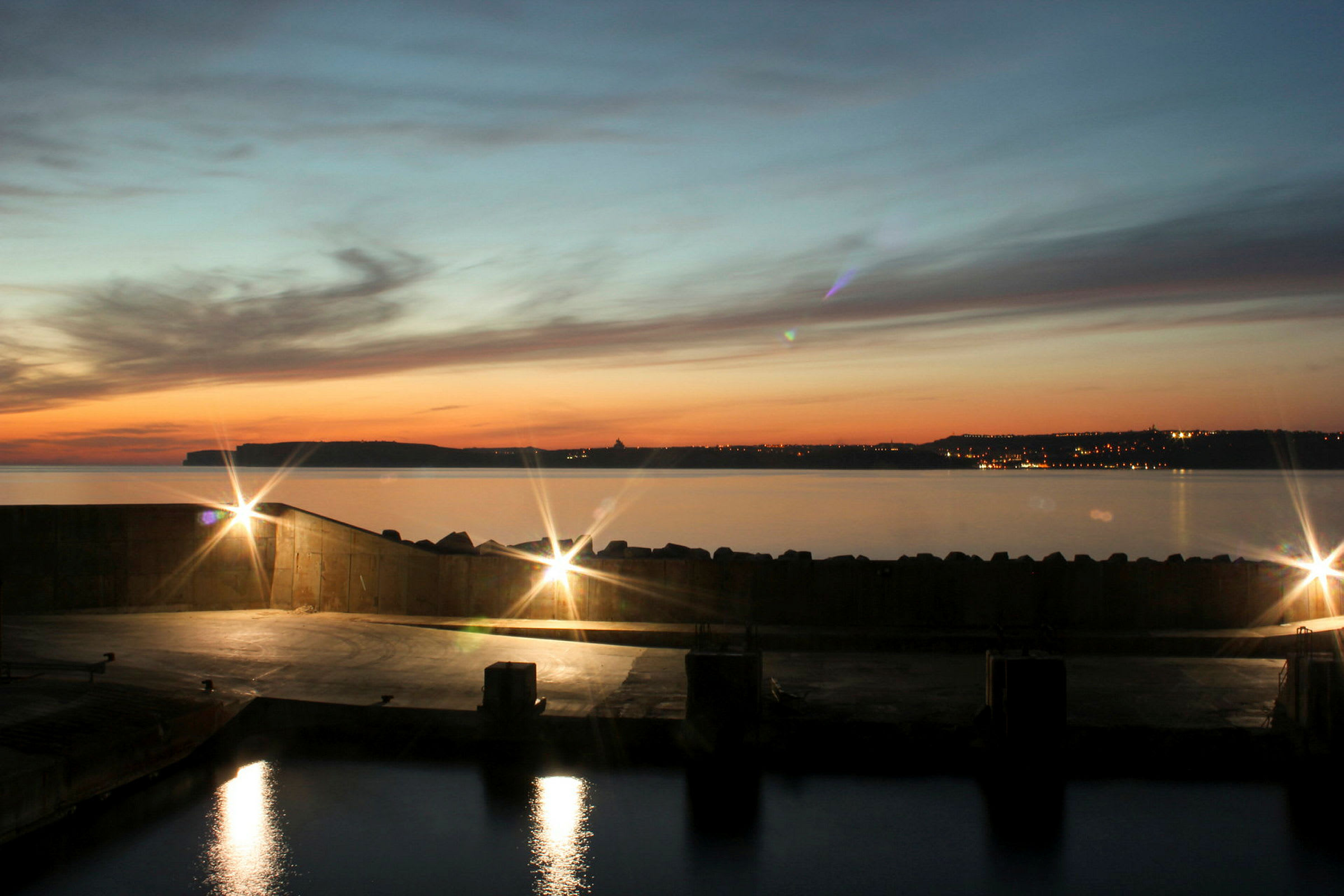 Havneområde med lyseffekter på vannet, kysten og bylysene er synlige ved solnedgang. (AI-generert tekst)