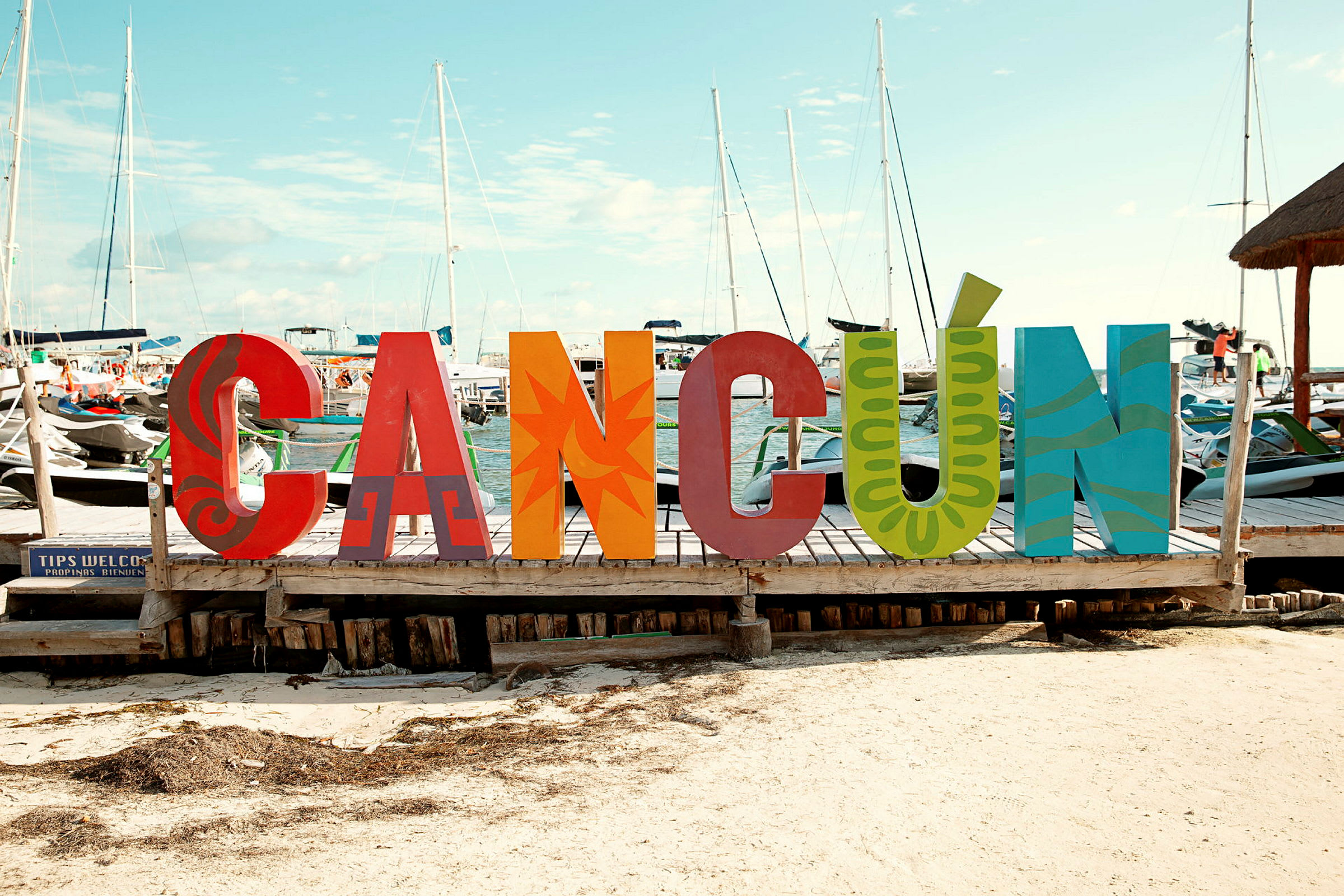 Fargerike bokstavsskulpturer som danner ordet 'Cancún' står på en brygge med båter og master i bakgrunnen. (AI-generert tekst)