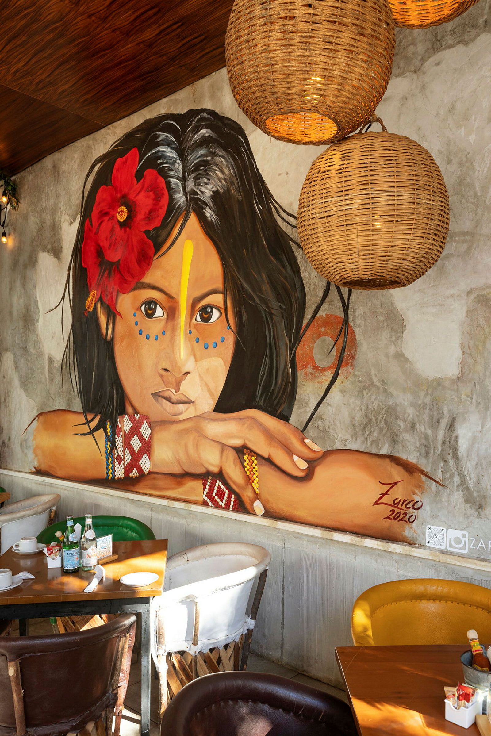 Et veggmaleri av en ung kvinne med en rød blomst i håret og fargerike mønstre, omringet av interiør i en restaurant. (AI-generert tekst)