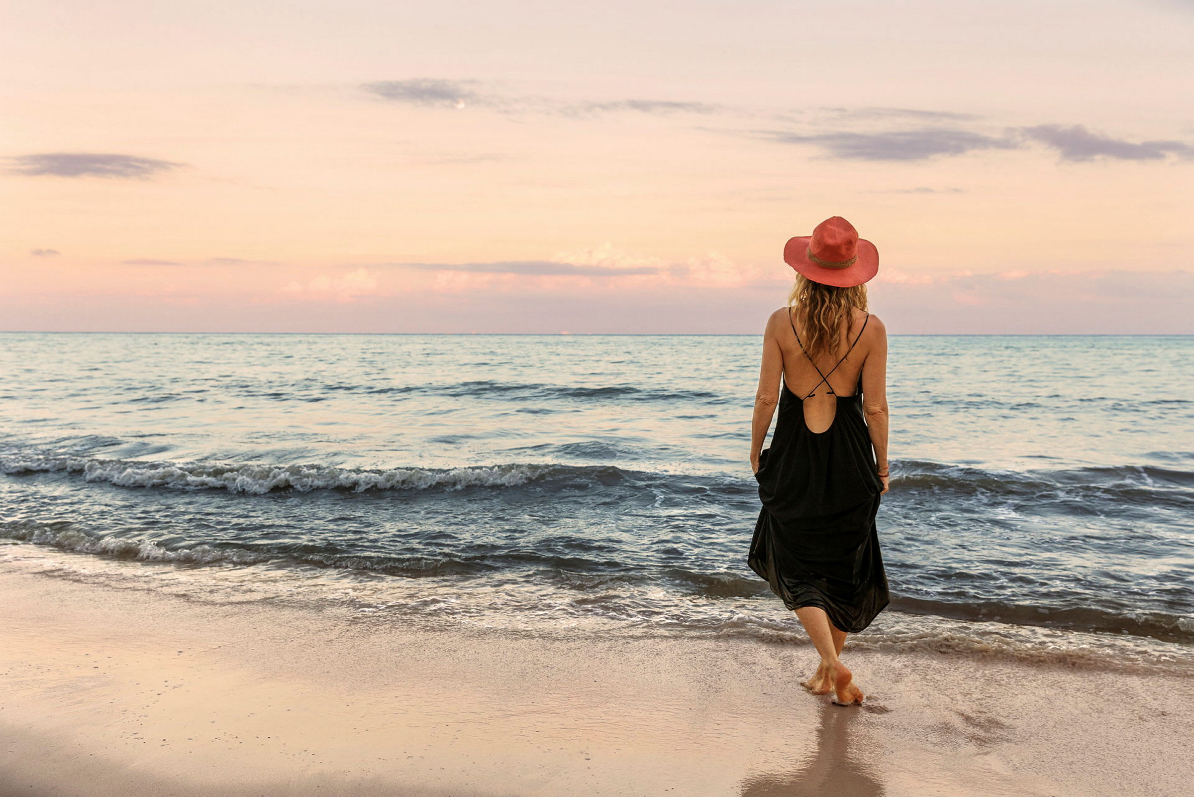 En person med en rød hatt og svart kjole går på en sandstrand ved havet under en pastellfarget himmel. (AI-generert tekst)