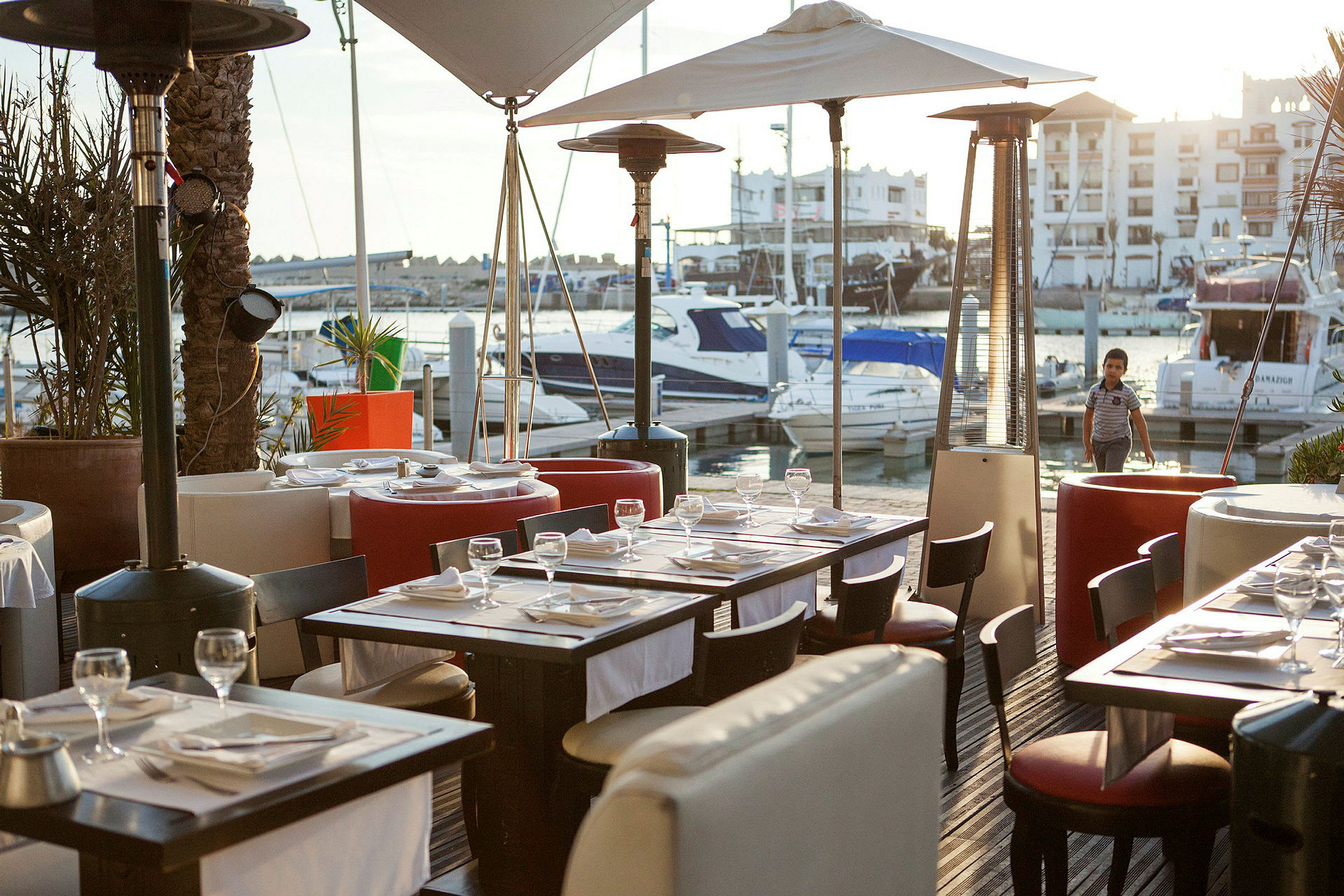 Utendørs restaurant ved en marina med båter og bygninger i bakgrunnen. (AI-generert tekst)