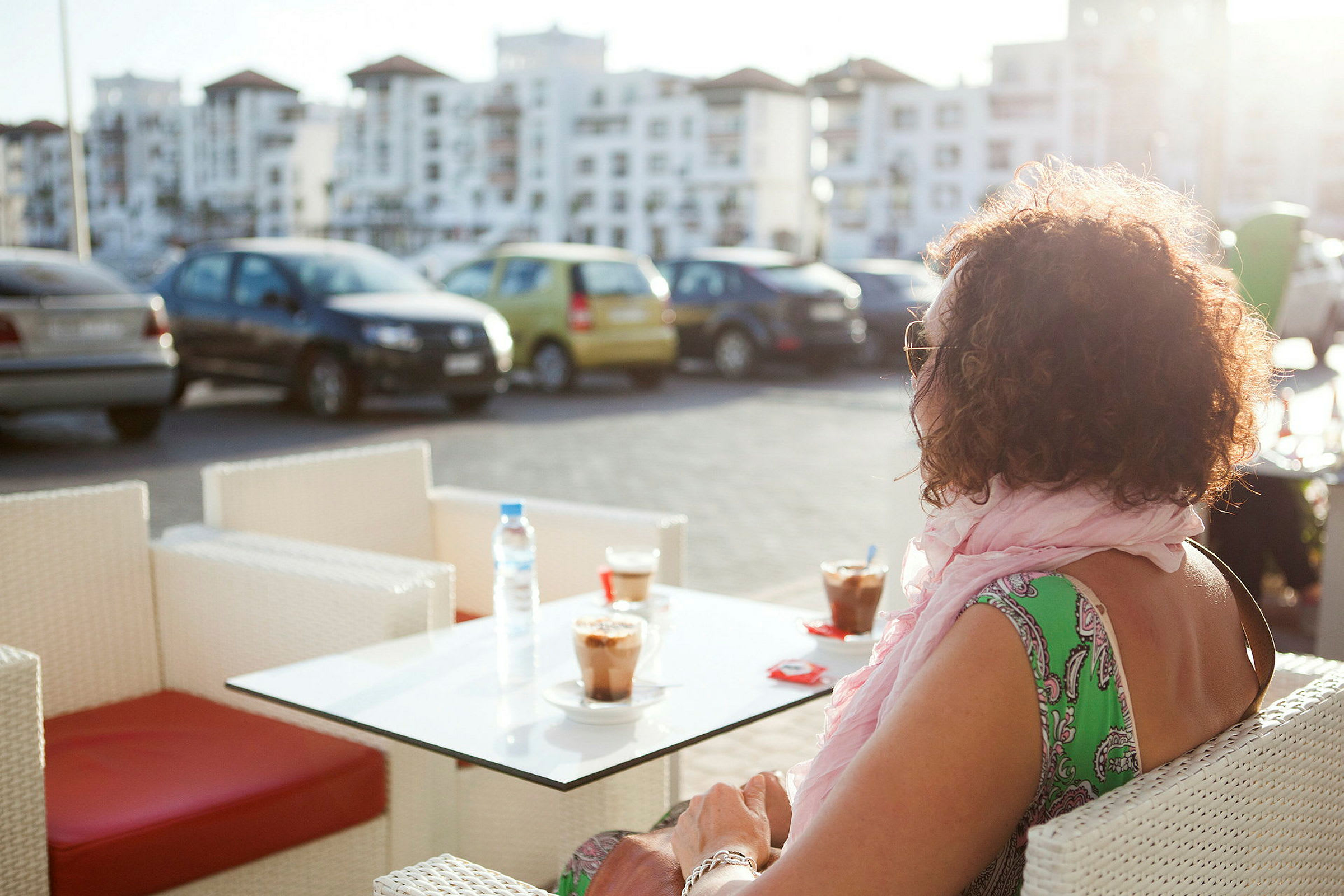 En person sitter ved et bord utendørs med to kaffedrikker, i bakgrunnen ses parkerte biler og bygninger. (AI-generert tekst)