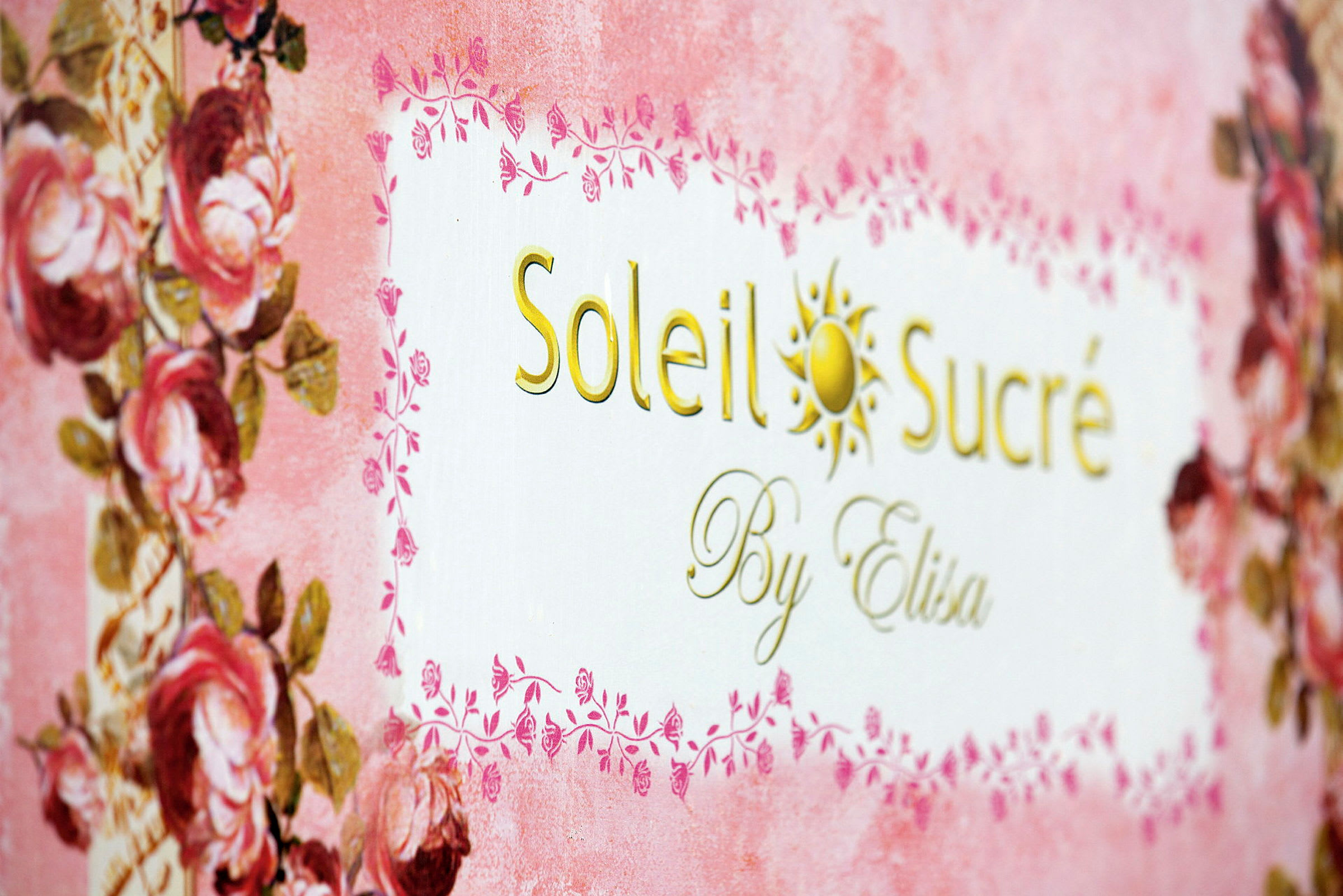 Teksten 'Soleil Sucré By Elisa' på en dekorert bakgrunn med rosa og blomsterdetaljer. (AI-generert tekst)