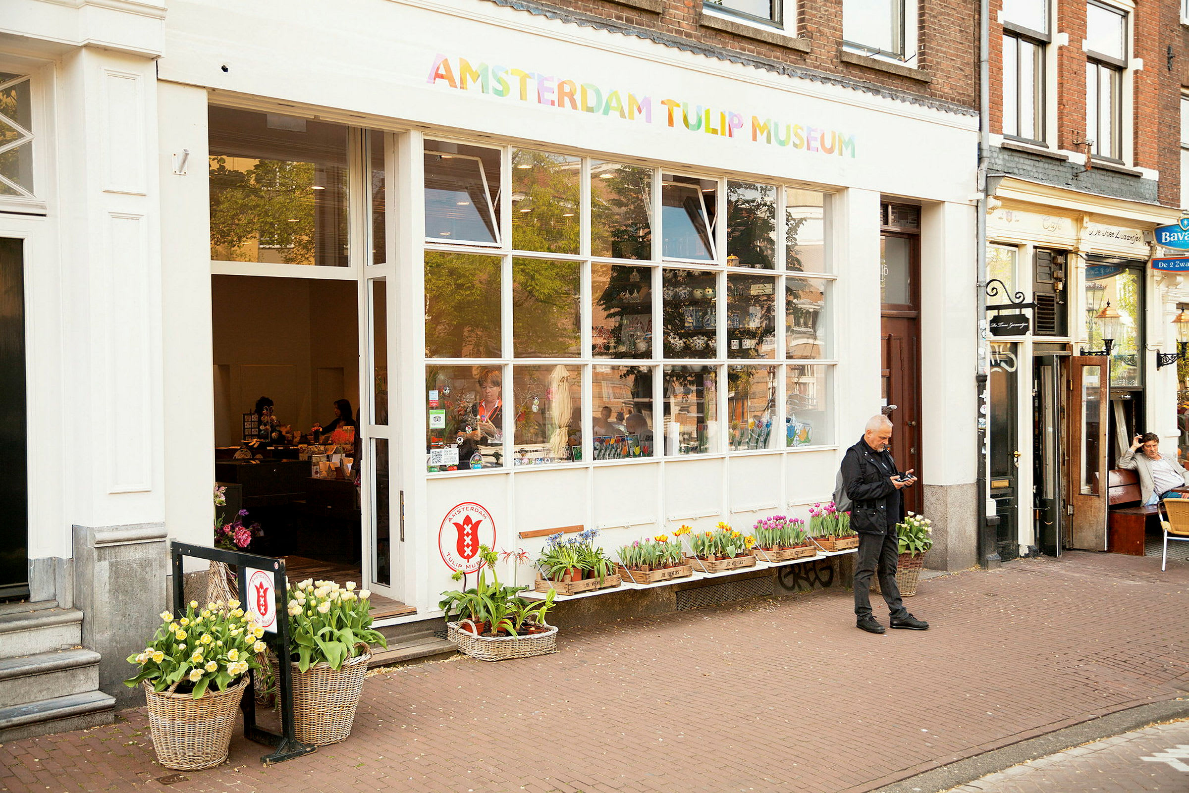 Inngangen til Amsterdam Tulip Museum, med blomster foran og mennesker inne i butikken. (AI-generert tekst)