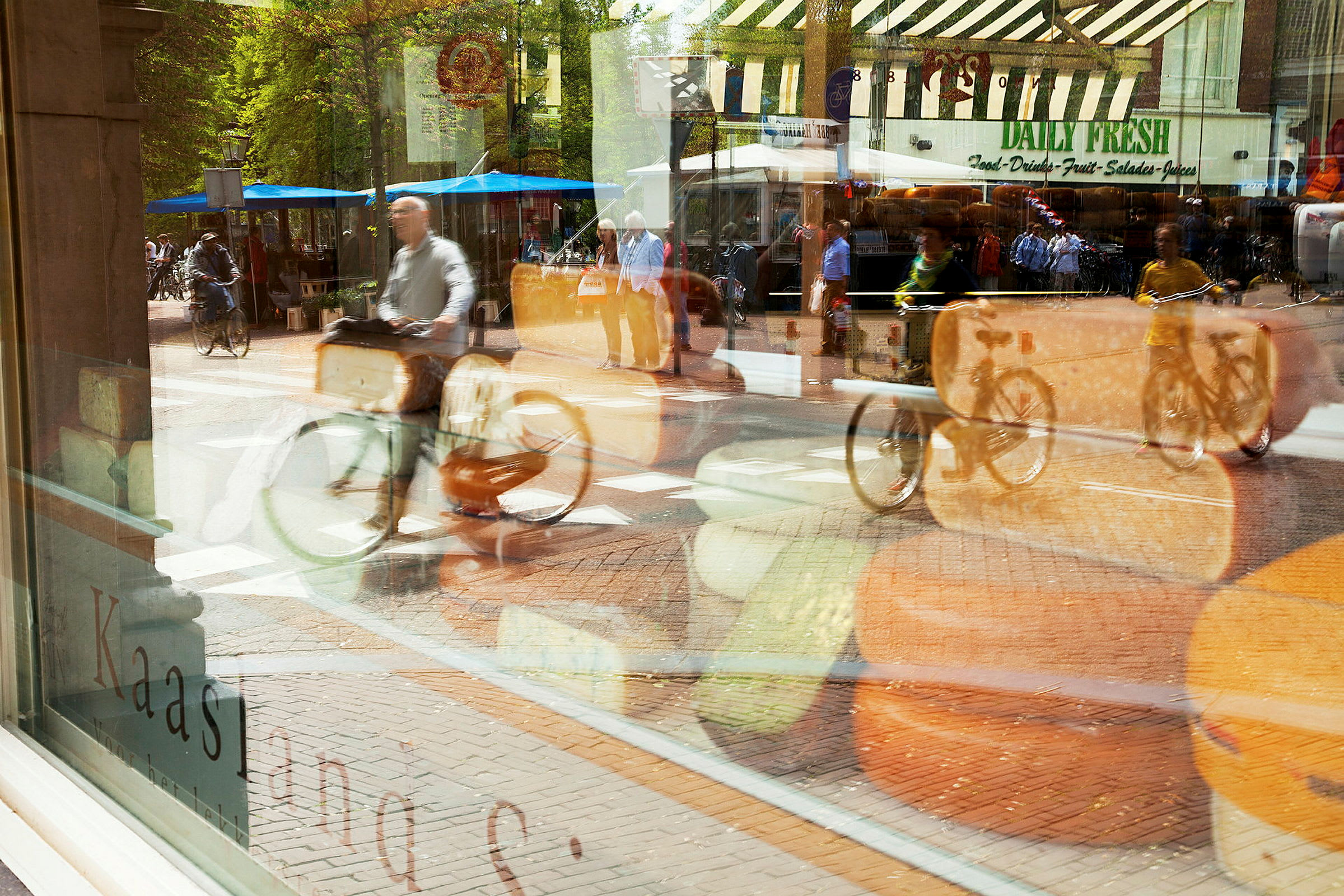 Et butikkvindu med ost og en refleksjon av en gate med syklister og markedsboder vises. (AI-generert tekst)