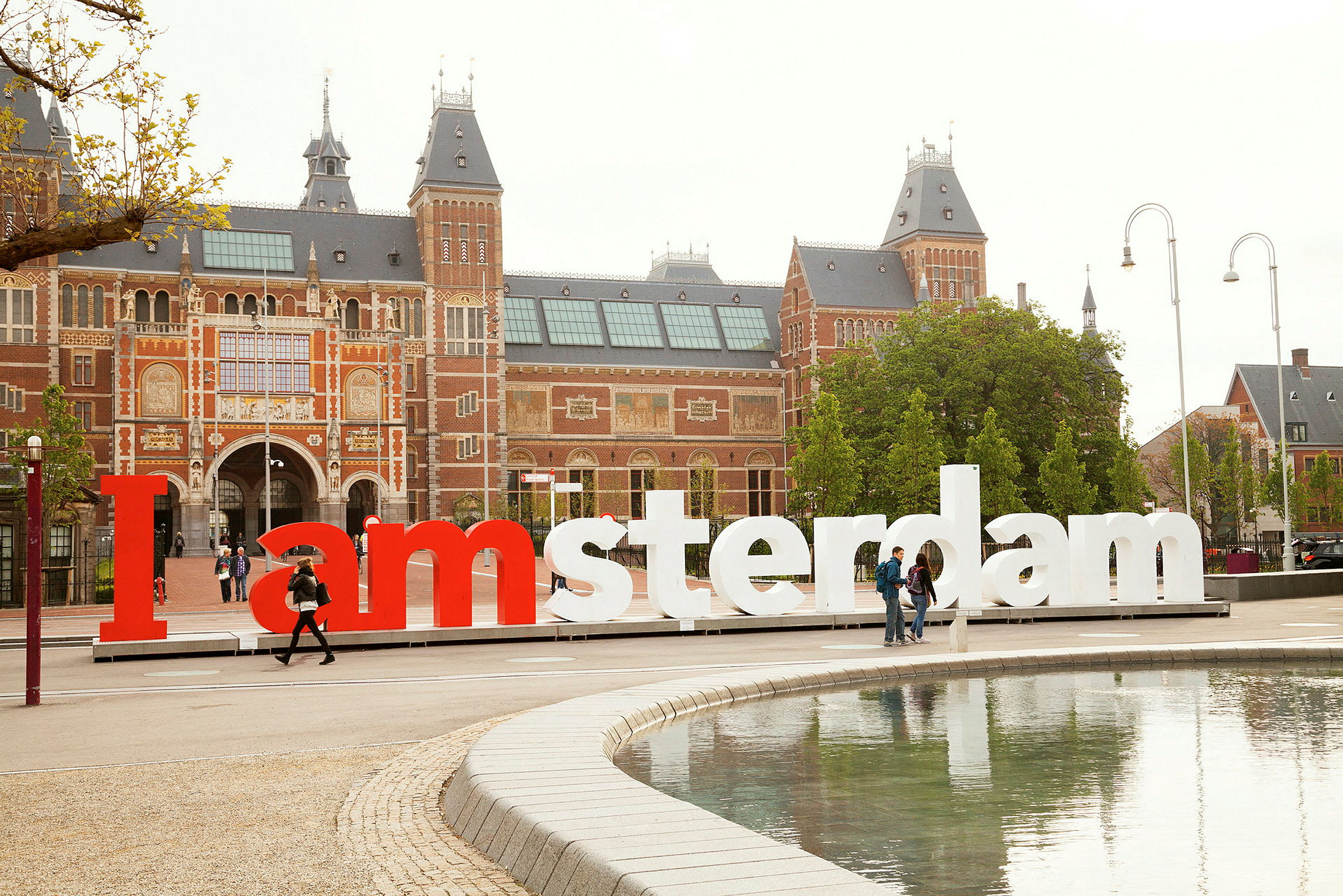 Stor bygning med murstein og flere tårn bak et I amsterdam-skilt, ved vannet. (AI-generert tekst)