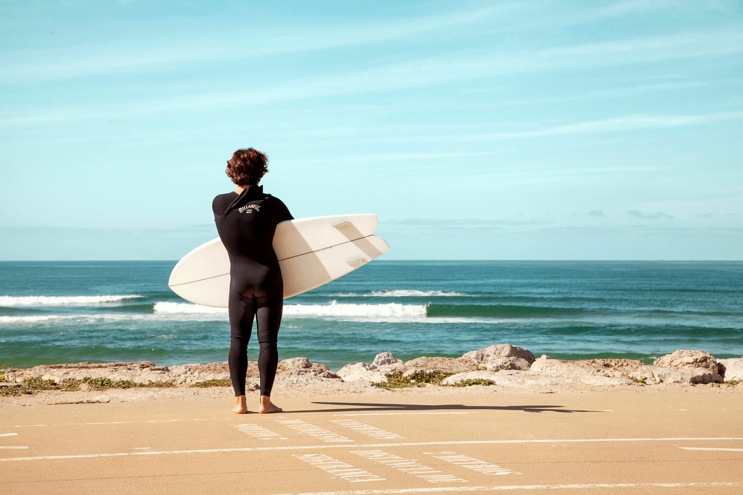 En person i våtdrakt holder et surfebrett og står vendt mot havet. (AI-generert tekst)