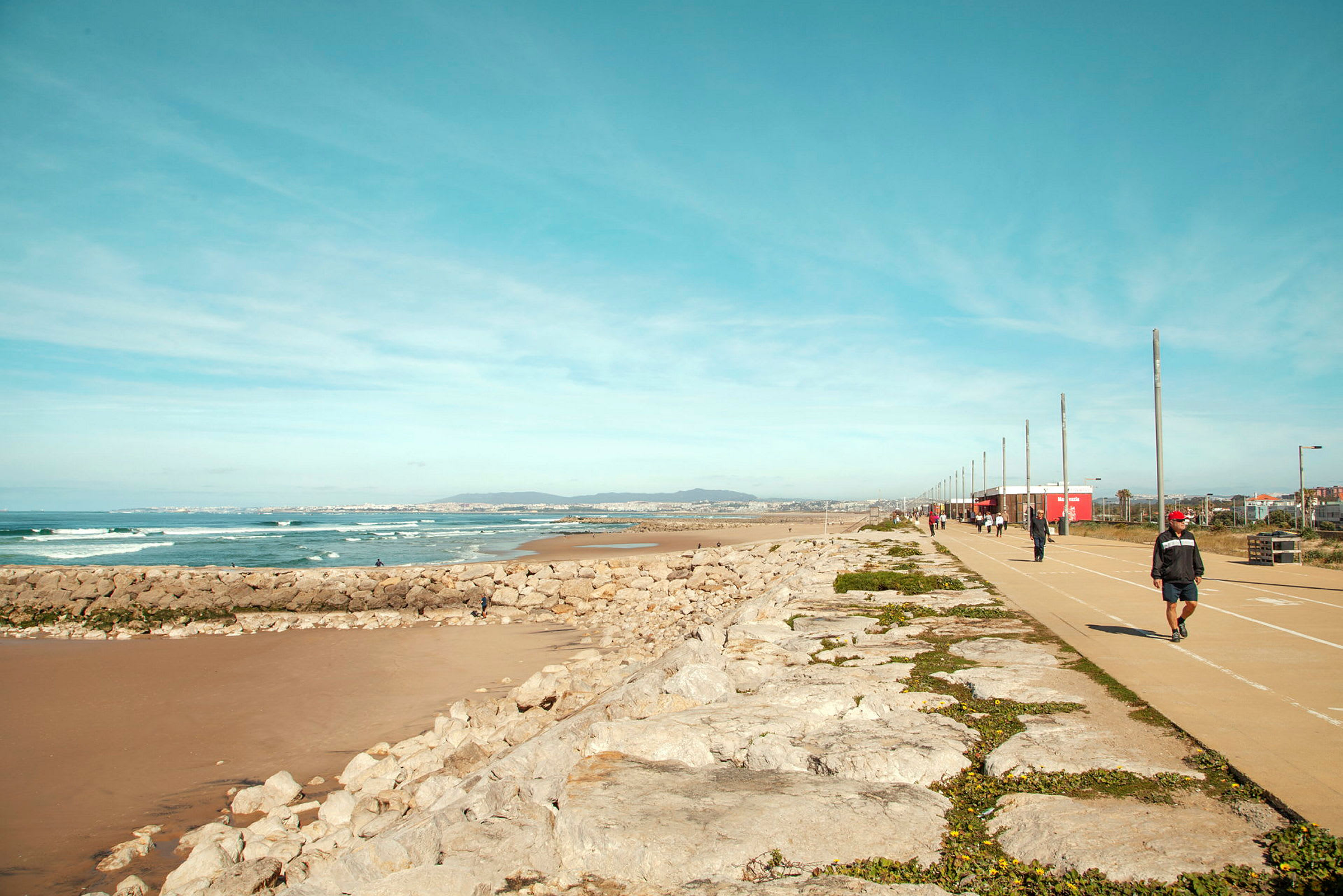 En strandpromenade med en gangsti ved siden av havet og klipper, mennesker går tur. (AI-generert tekst)