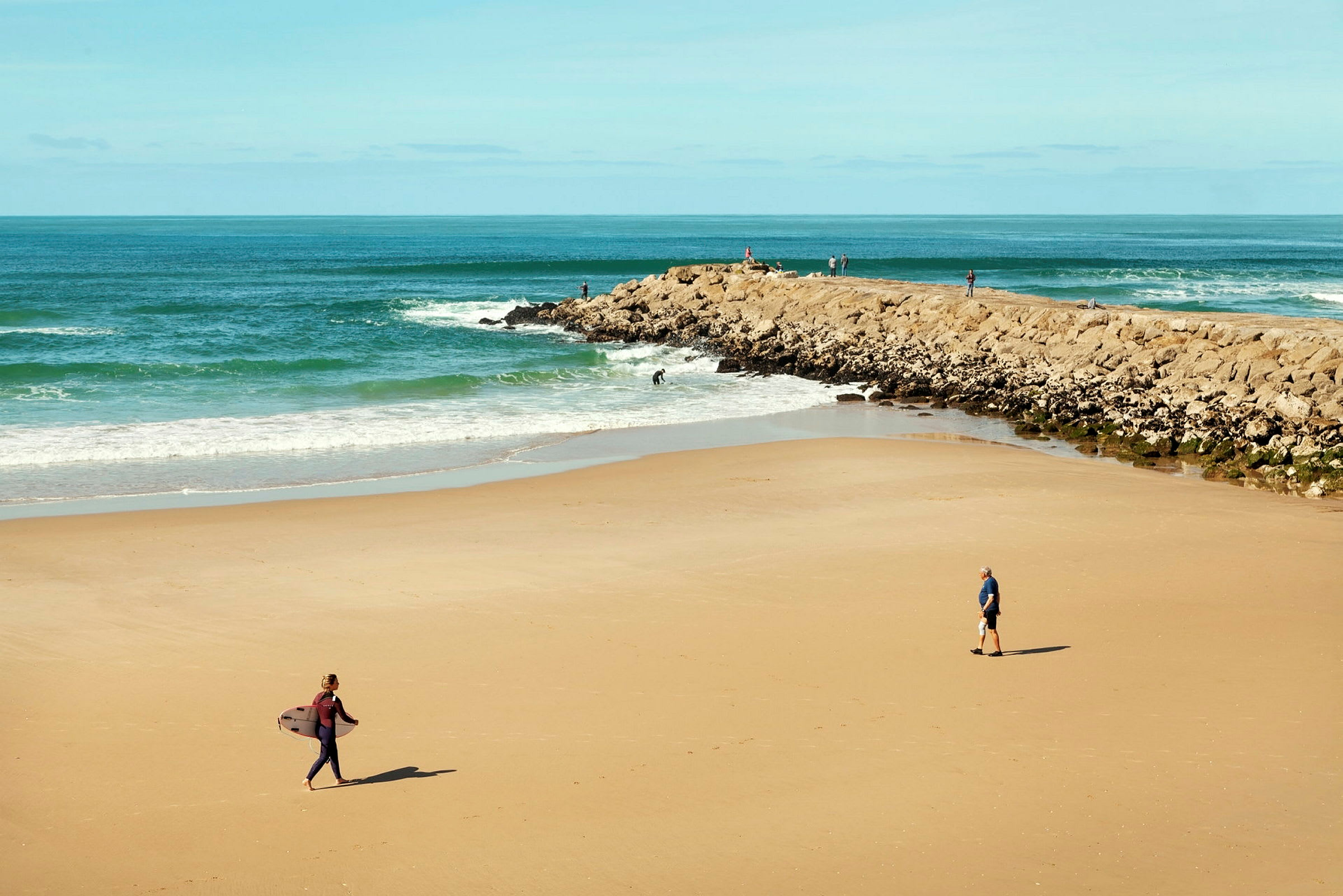 En strand med noen personer, en av dem bærer et surfebrett. (AI-generert tekst)