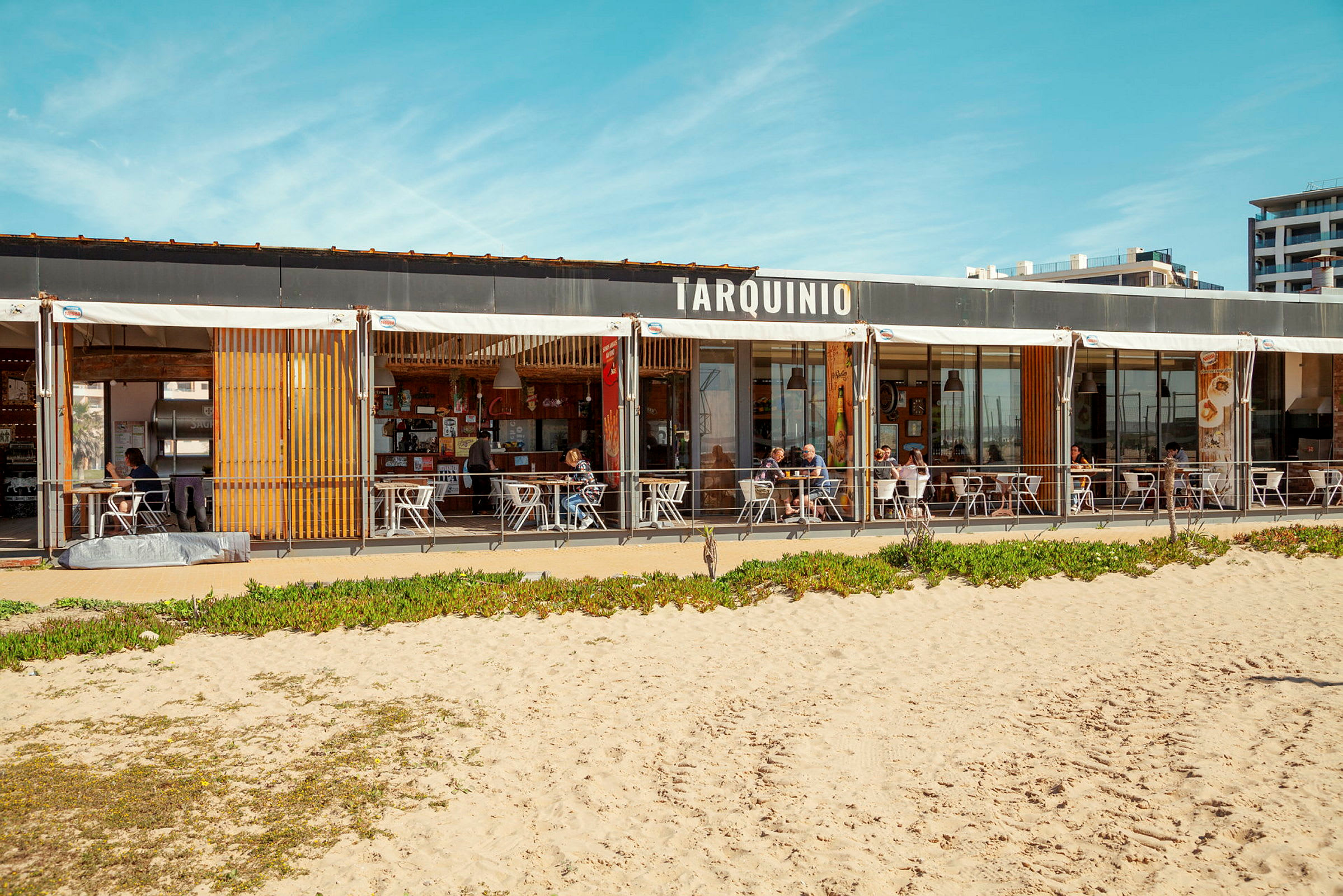 En strandrestaurant med skilt som sier 'Tarquinio', utemøbler og sandstrand i forgrunnen. (AI-generert tekst)