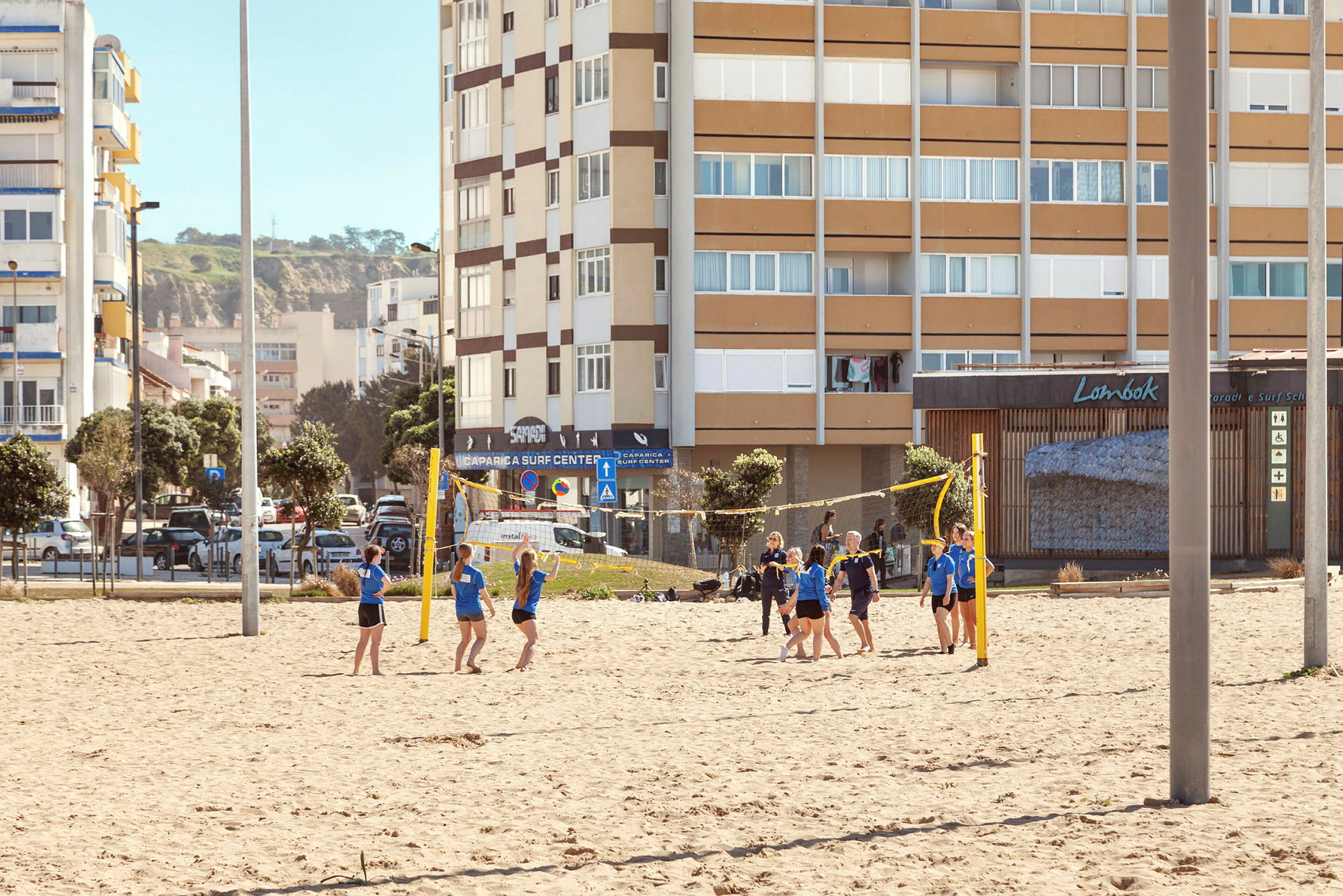 En gruppe mennesker spiller volleyball på en sandbane med bygninger i bakgrunnen. (AI-generert tekst)