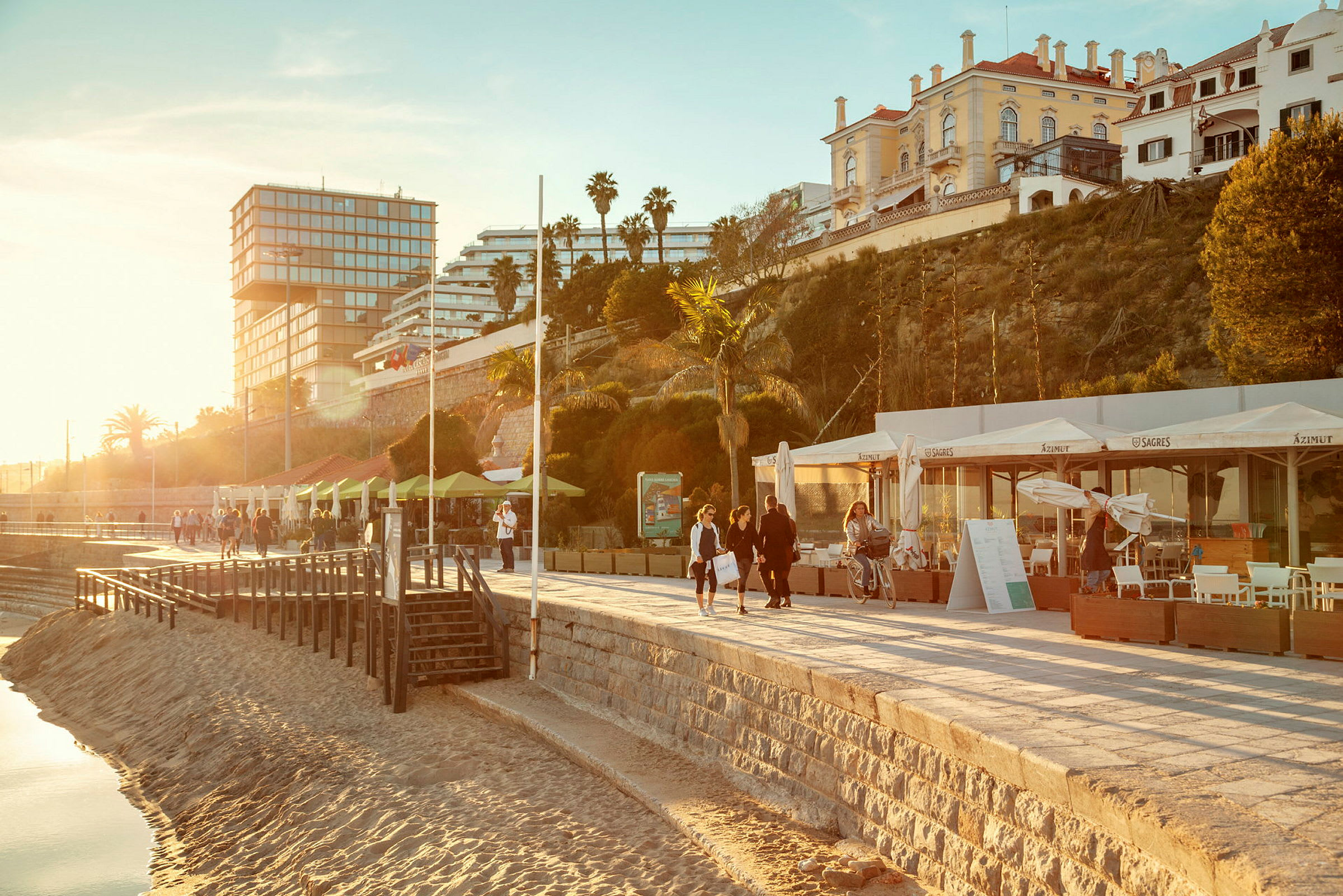 Strandpromenade med bygninger i bakgrunnen og mennesker som går tur. (AI-generert tekst)
