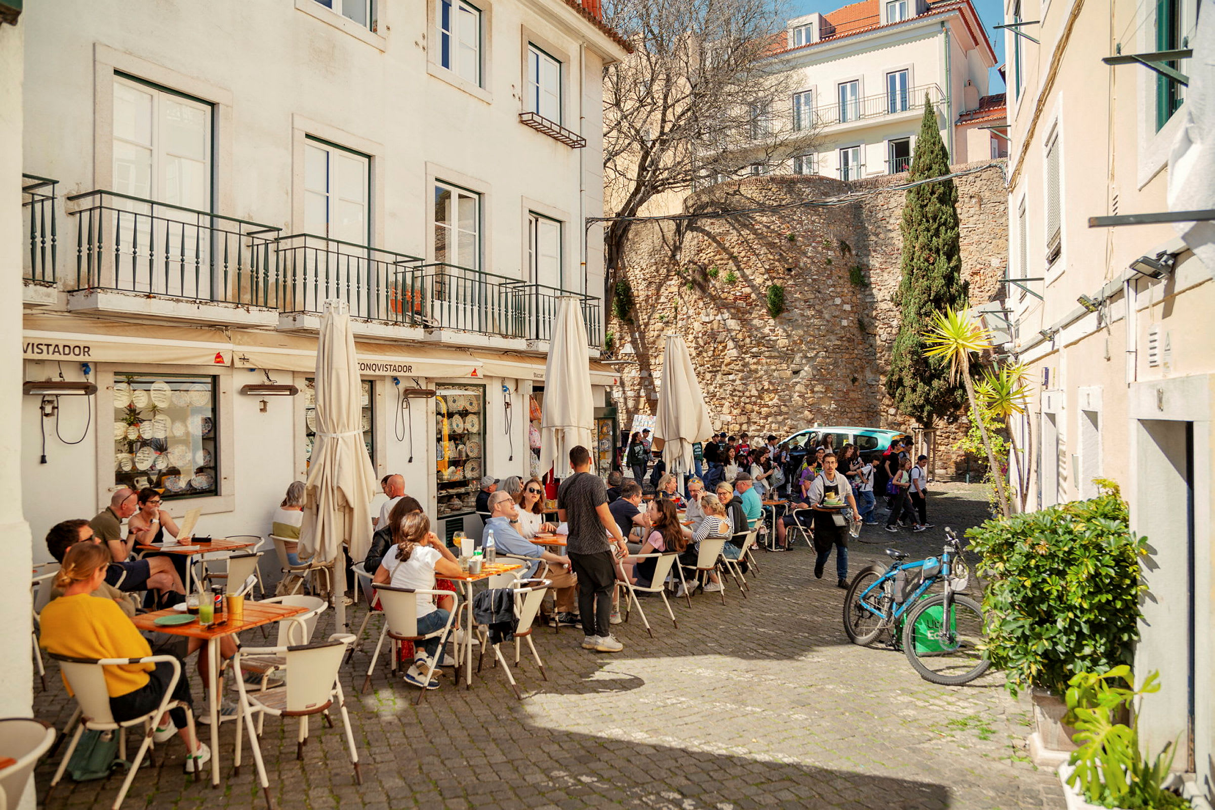 En utendørsterrasse på en restaurant med flere mennesker som sitter ved bord og koser seg med måltidet sitt. I bakgrunnen sees en steinmur og en trekledd bygning. (AI-generert tekst)