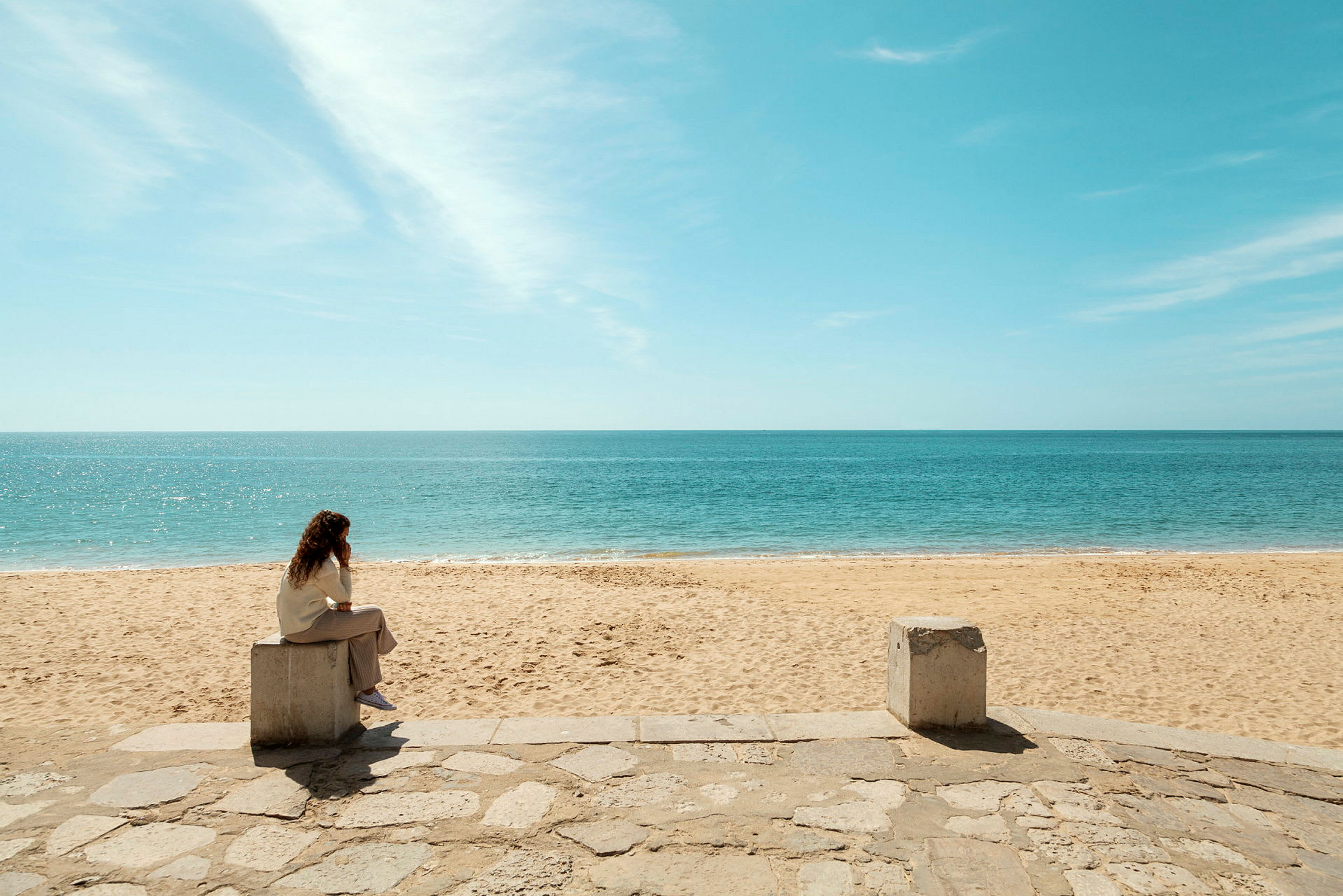 En person sitter på en betongblokk ved en sandstrand og ser utover havet. (AI-generert tekst)