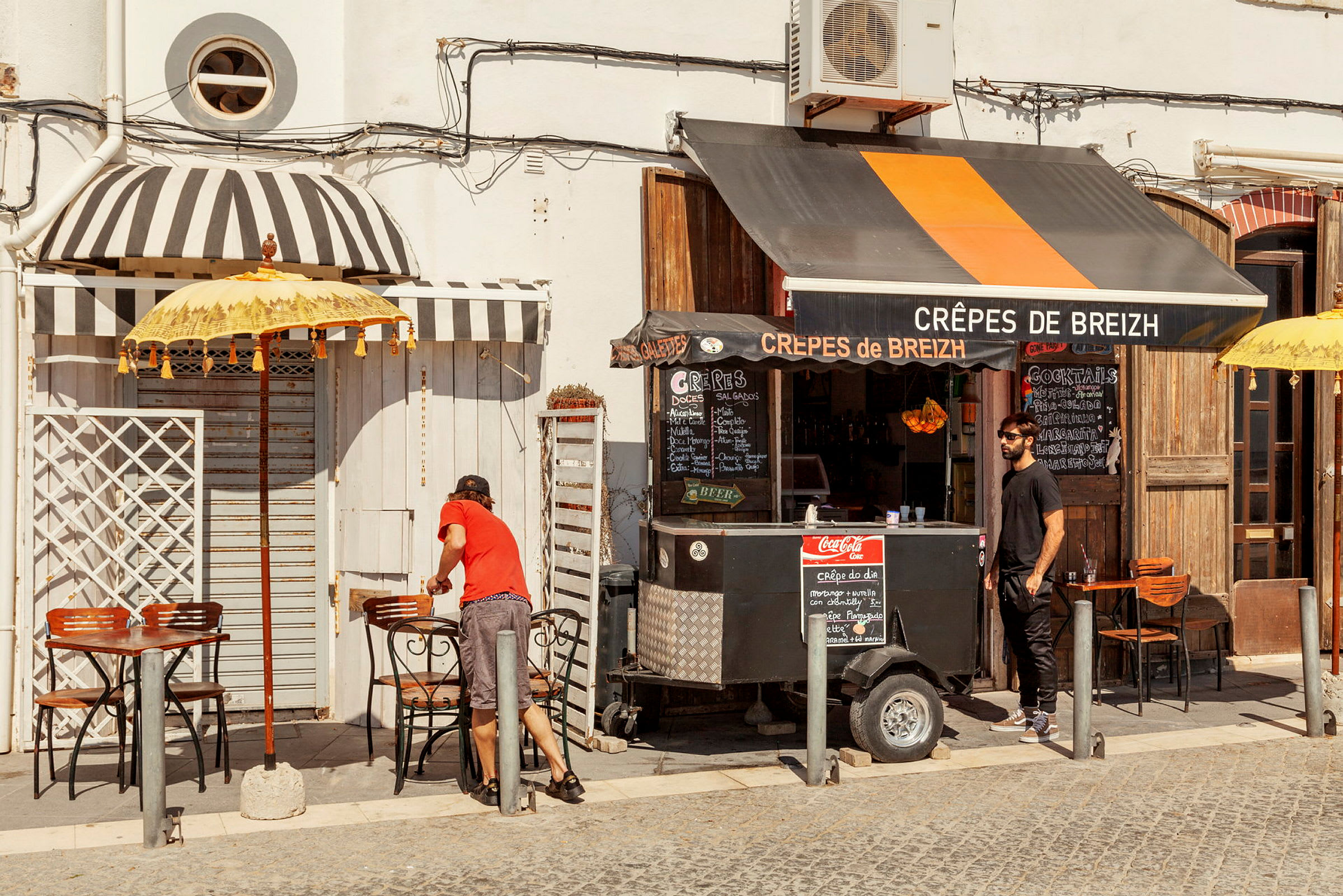 En liten matvogn som selger crêpes med bord og sitteplasser utendørs på en solrik dag. (AI-generert tekst)