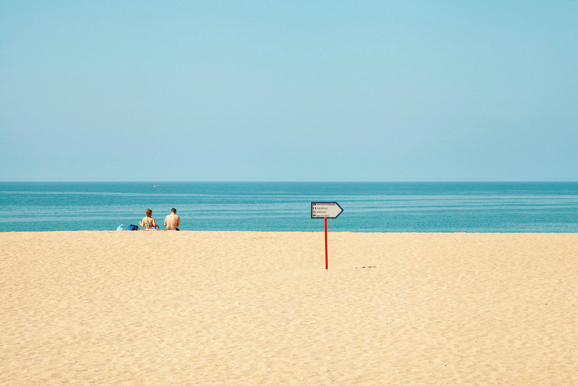 To personer sitter på en sandstrand med havet i bakgrunnen, og et veiskilt er synlig på stranden. (AI-generert tekst)