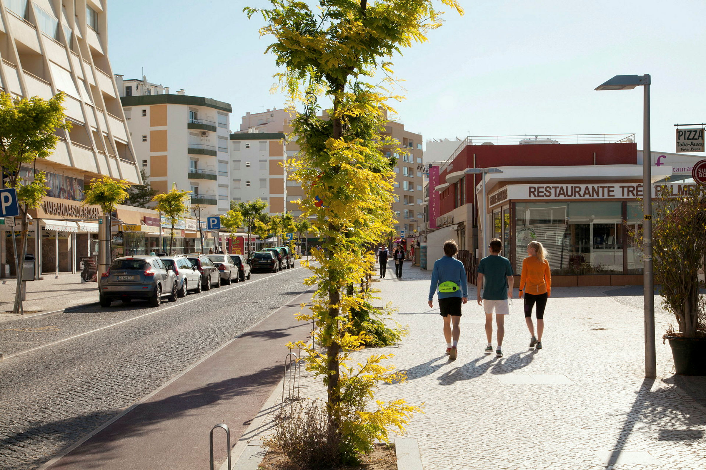 Personer går langs en gate med parkerte biler og bygninger i bakgrunnen. (AI-generert tekst)