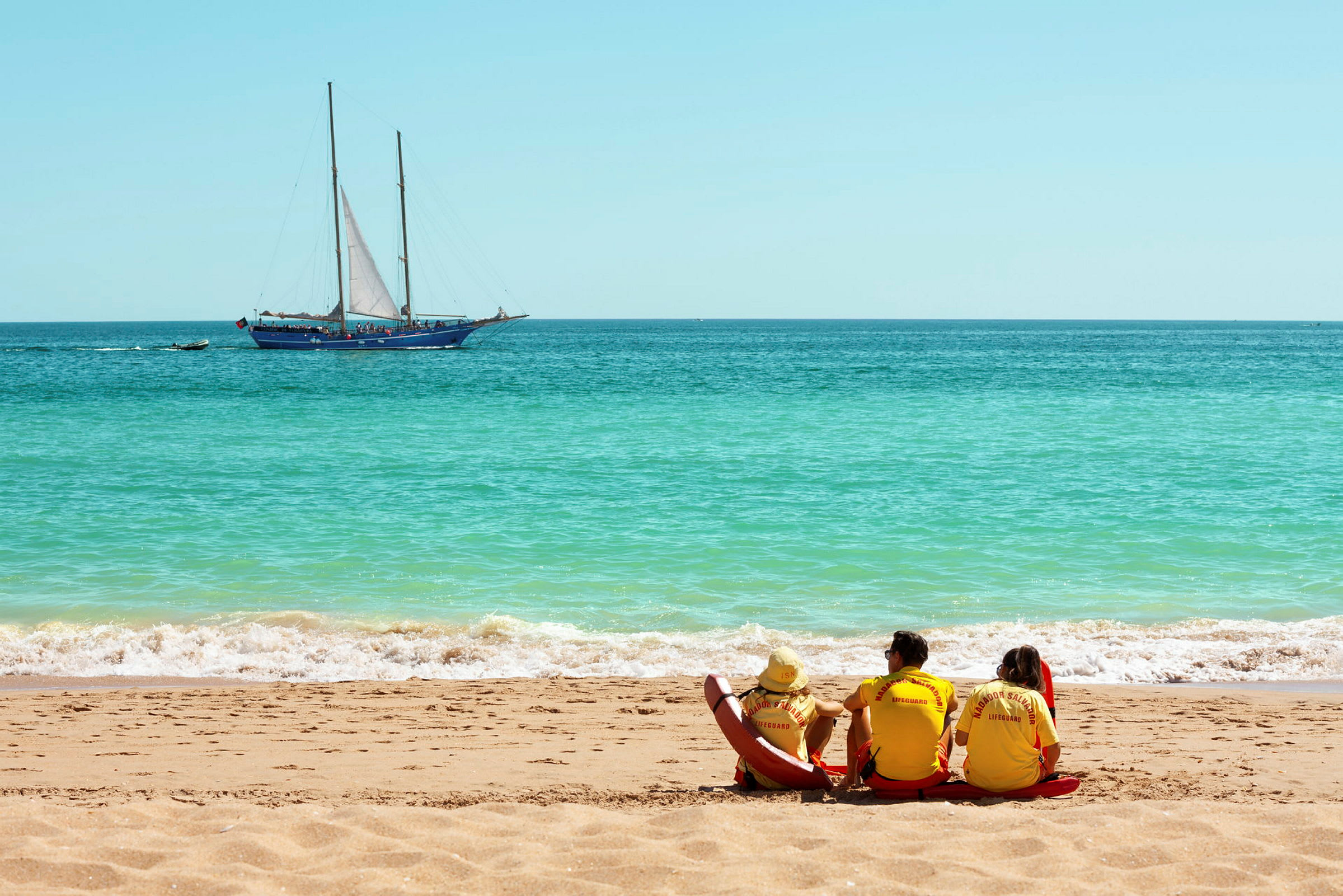 Livredderne sitter på en strand med en seilbåt på havet i bakgrunnen. (AI-generert tekst)
