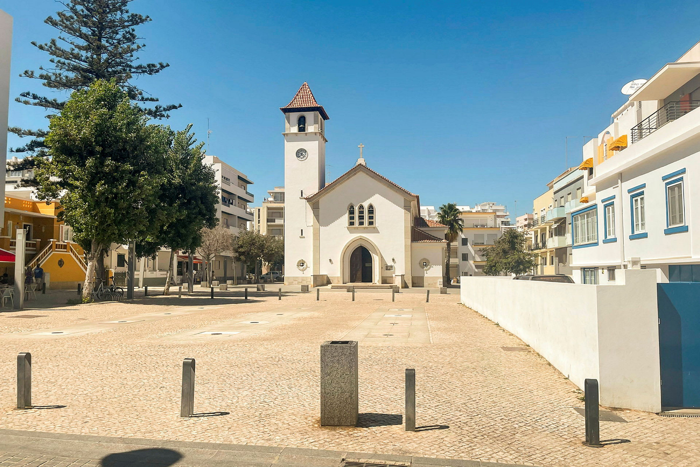 En hvit kirke med et klokketårn vises på et torg omgitt av bygninger. (AI-generert tekst)