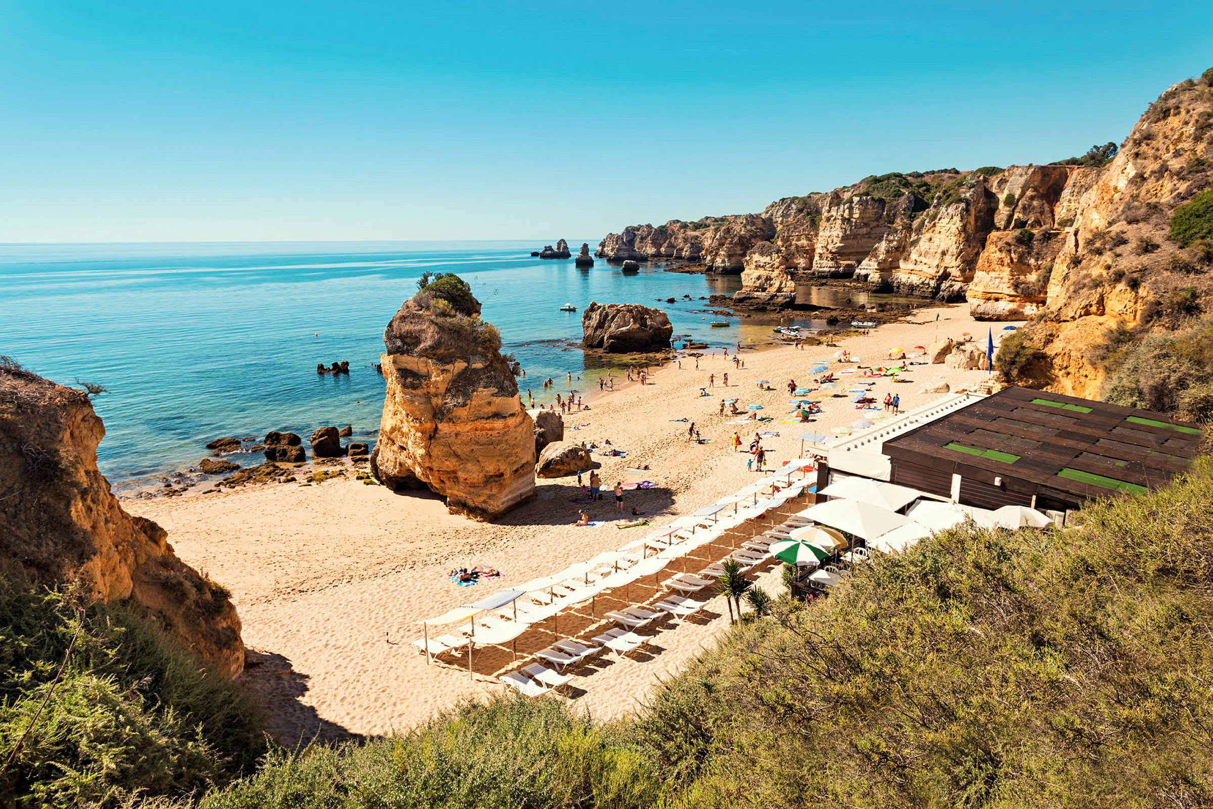 En sandstrand med solsenger og parasoller, omgitt av klippeformasjoner og et stille hav. (AI-generert tekst)