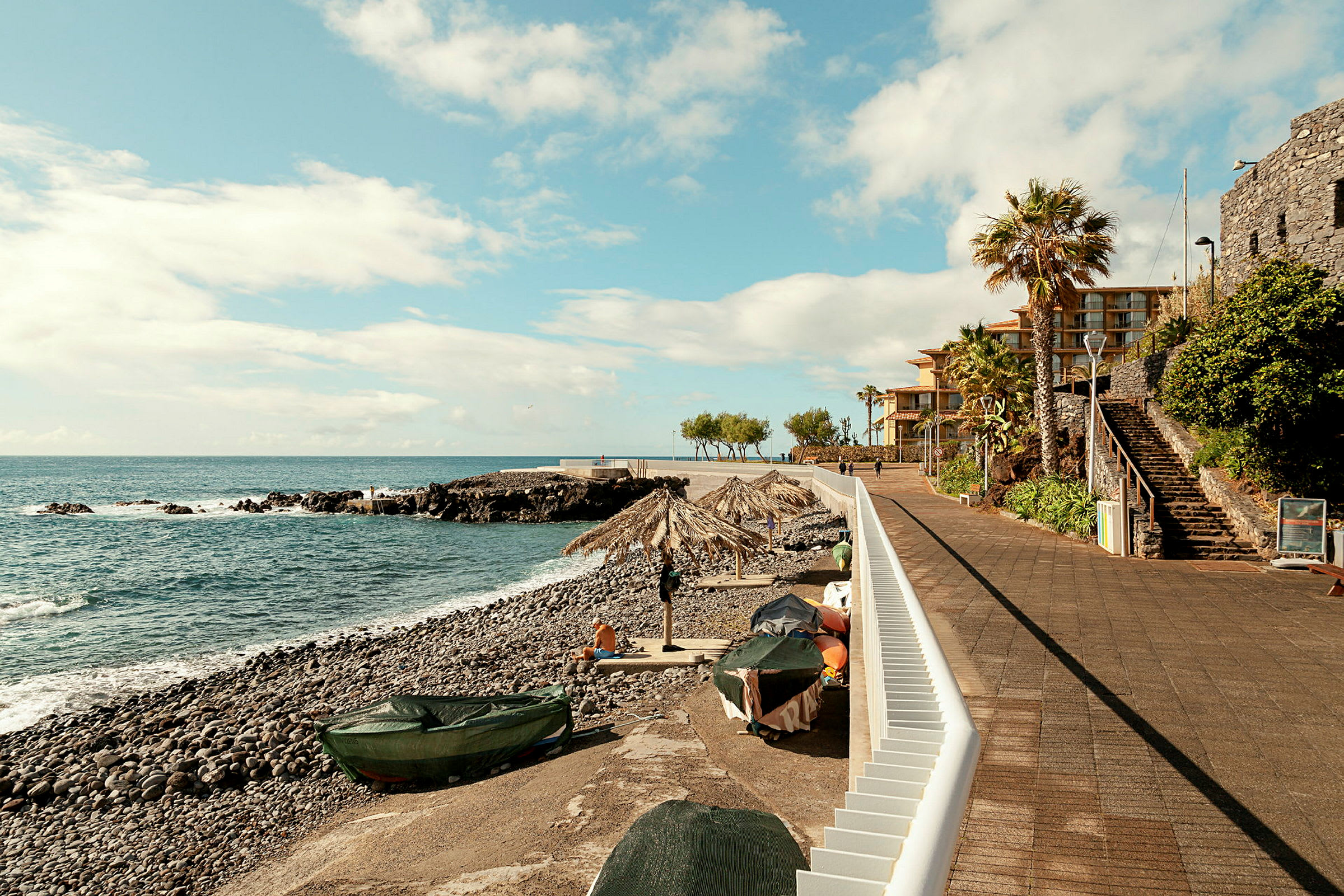 Utsikt over en strand med steinete kyst, båter og parasoller, samt en promenade med bygninger og palmer. (AI-generert tekst)