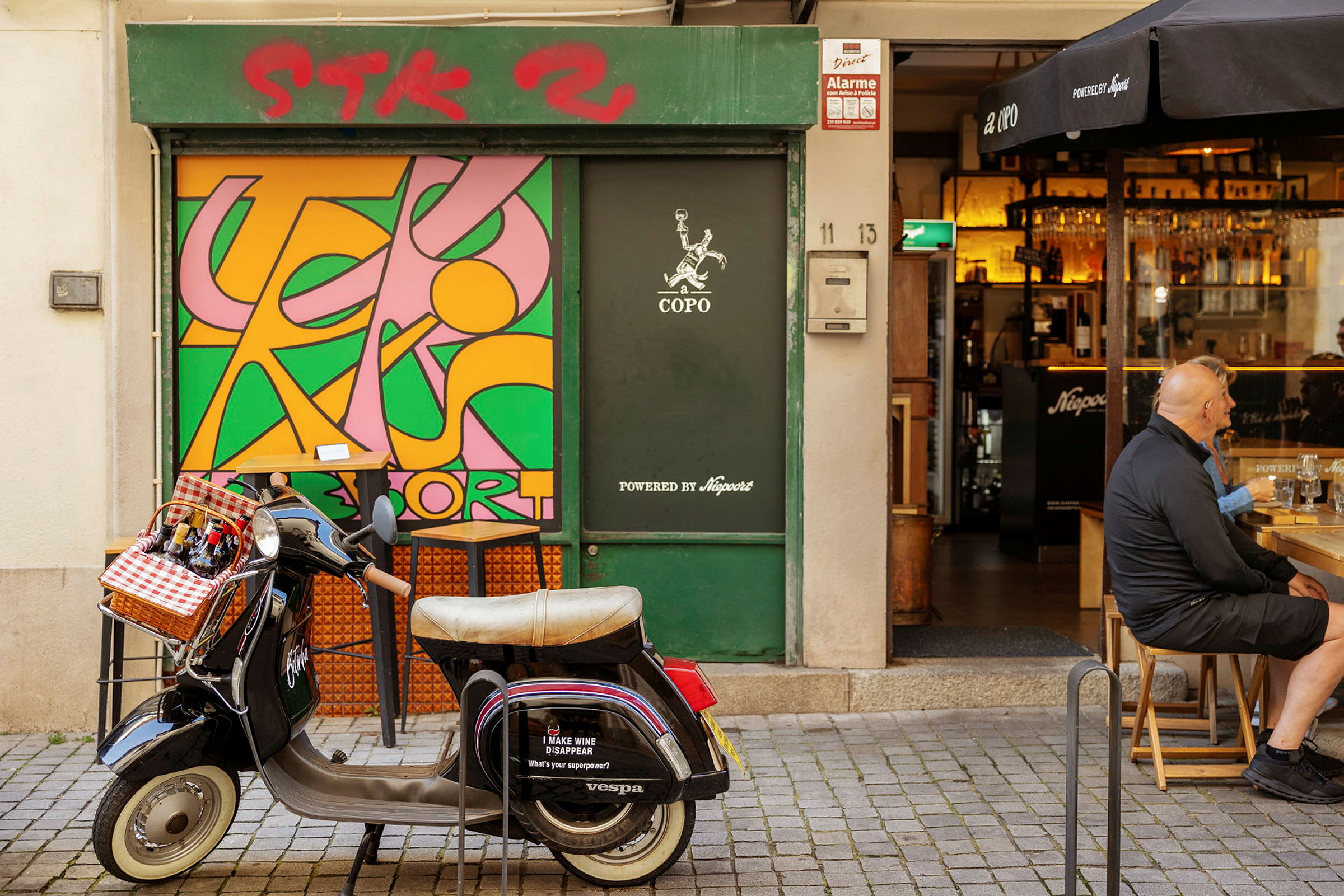 En Vespa med en kurv foran som inneholder flasker er parkert foran en butikk med et fargerikt veggmaleri. (AI-generert tekst)