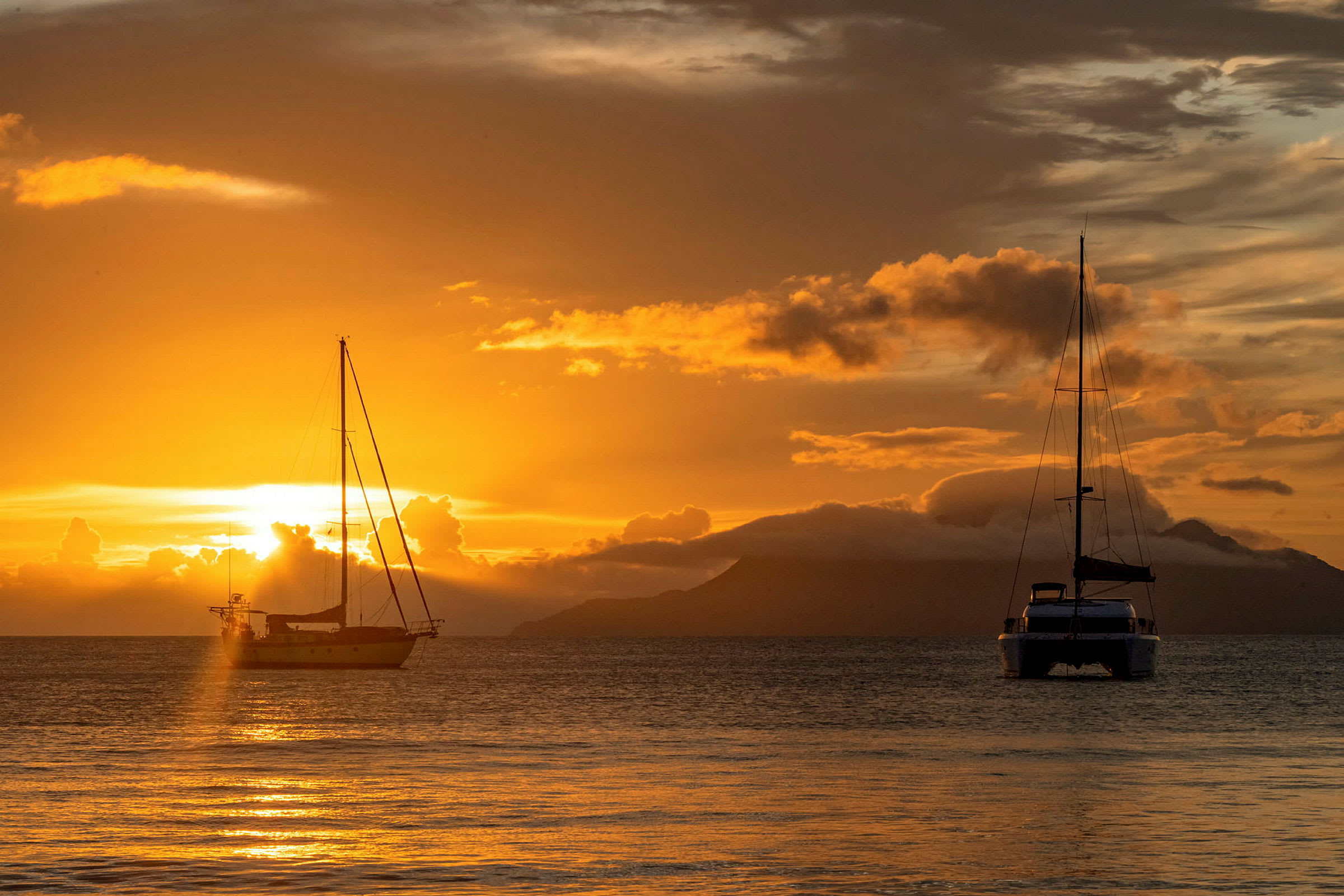 To seilbåter ligger forankret på havet under en solnedgang, hvor himmelen er fylt med gyldne og oransje toner. (AI-generert tekst)