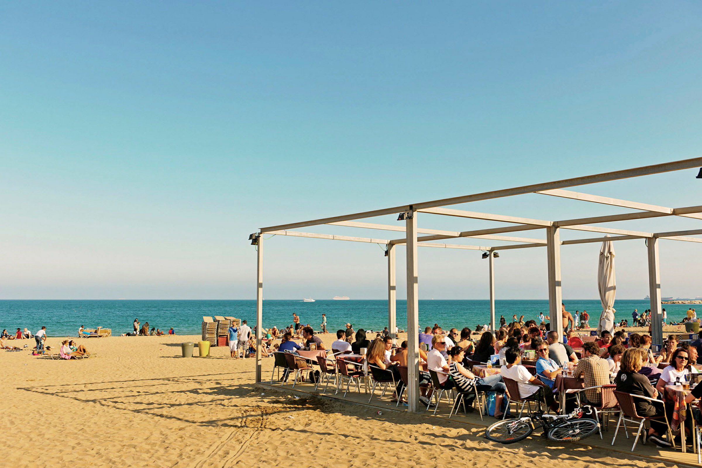 En strand med folk som sitter ved et kaféområde under et tak og andre som nyter stranden og havet. (AI-generert tekst)