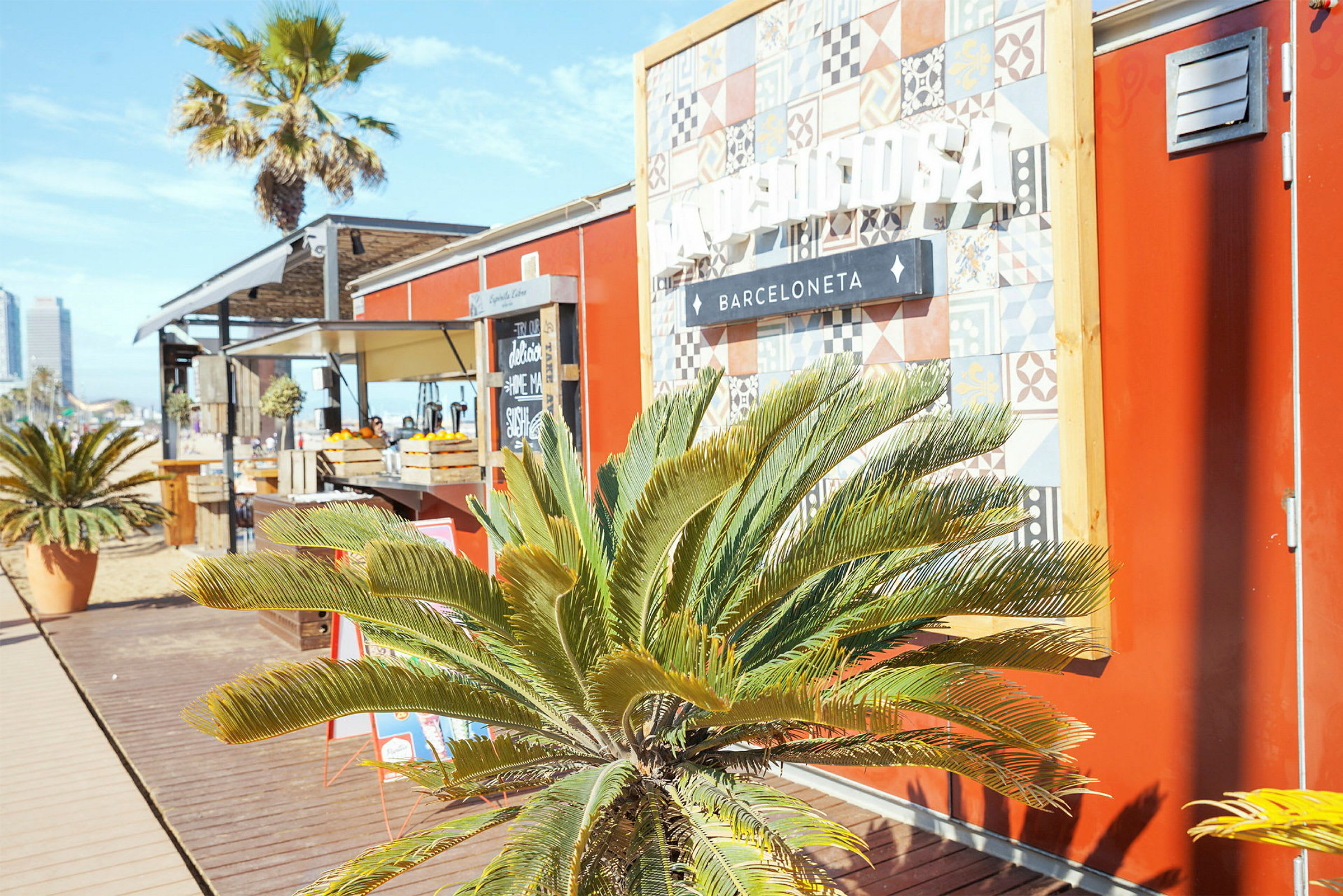En liten strandbar med røde vegger, et dekorativt skilt og grønne palmeplanter foran. (AI-generert tekst)