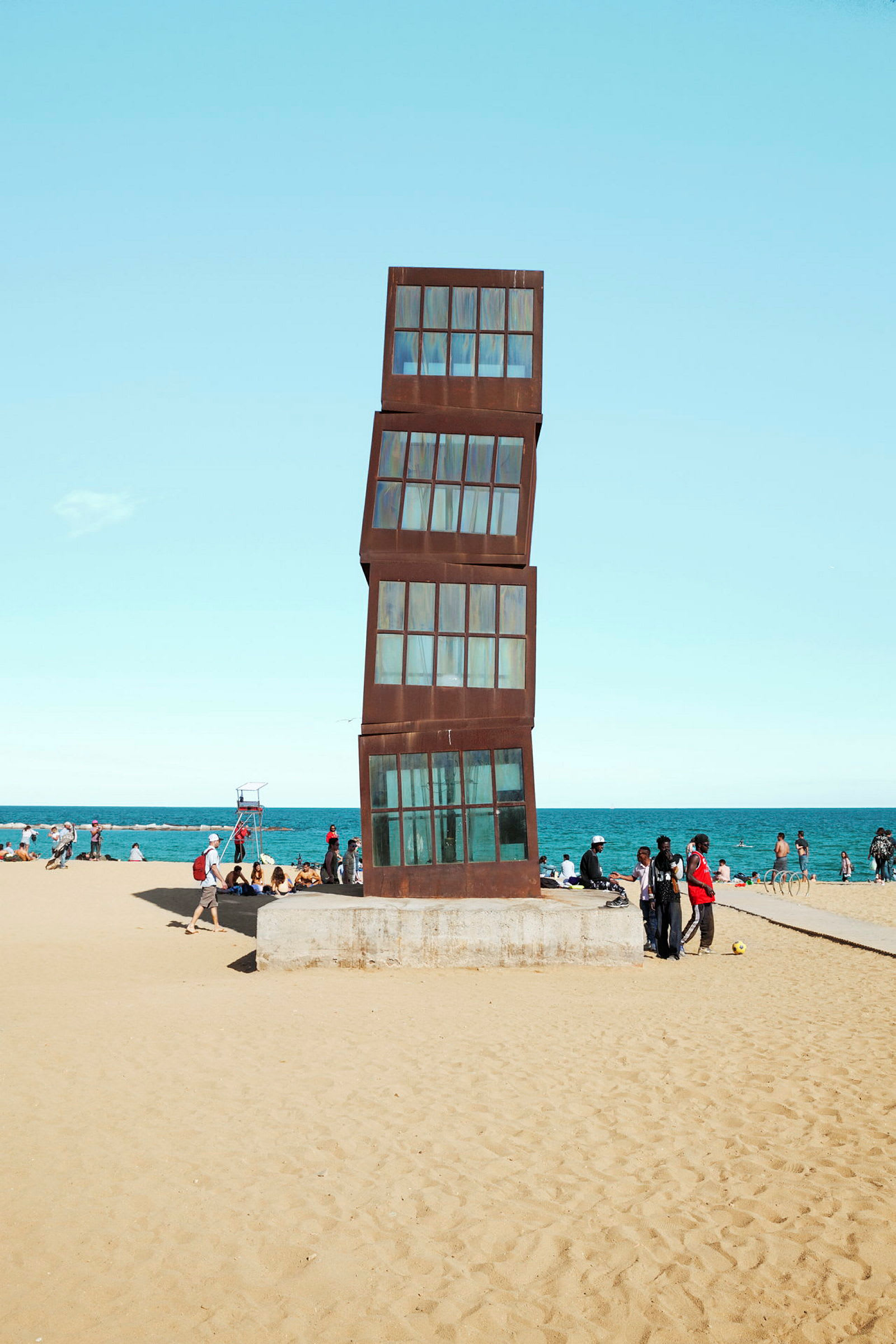En skrånende skulptur av stablede glass- og metallvinduer på en strand med havet i bakgrunnen. (AI-generert tekst)