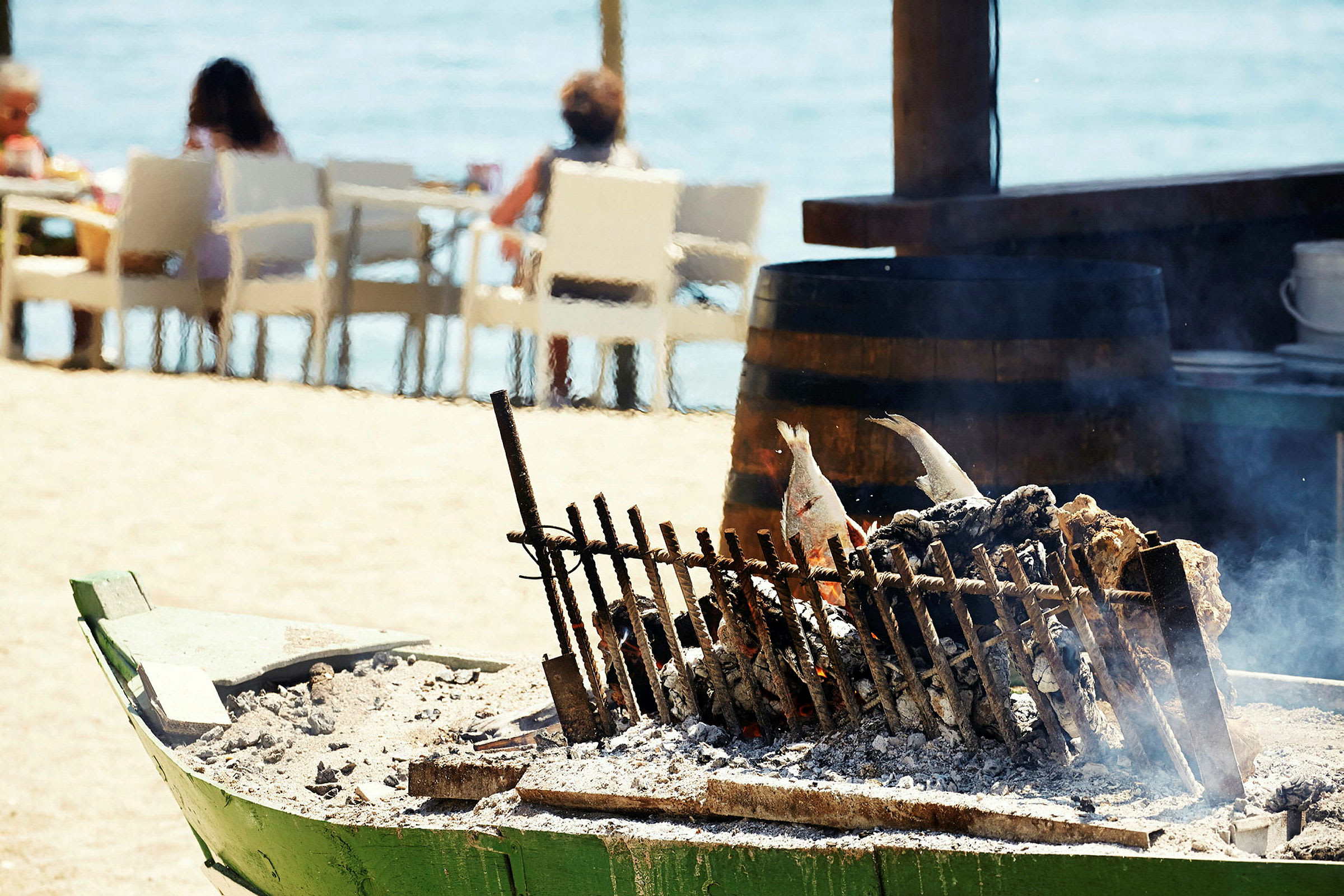 Grillet fisk på et spidd over glør, med personer sittende ved strandbord i bakgrunnen. (AI-generert tekst)