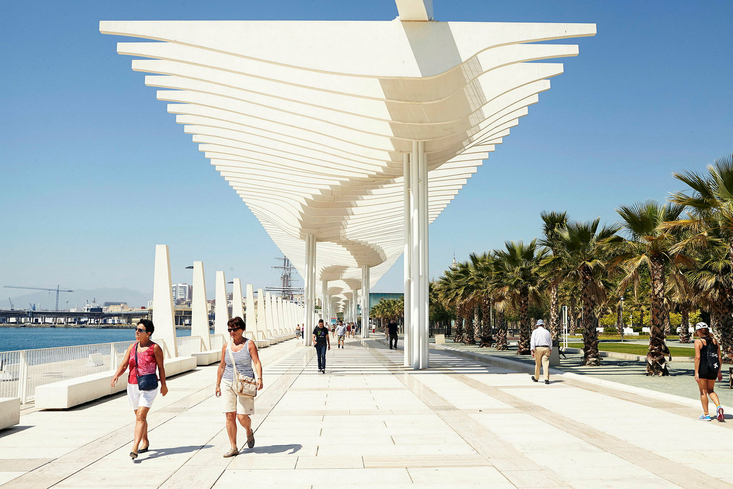 En strandpromenade med moderne arkitektur og palmer på en solfylt dag. Folk spaserer langs gangveien. (AI-generert tekst)