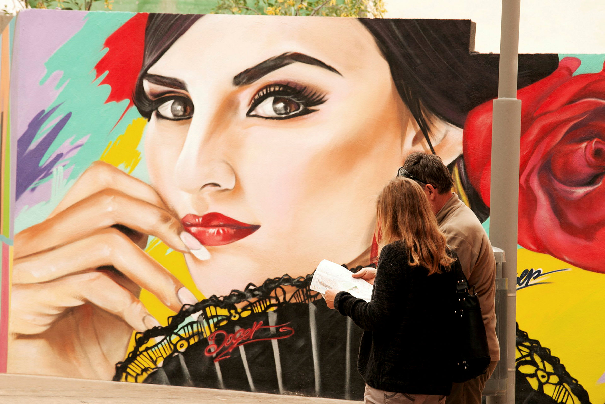 To personer står foran en vegg med fargerik graffitikunst som viser et ansikt og en rose. (AI-generert tekst)