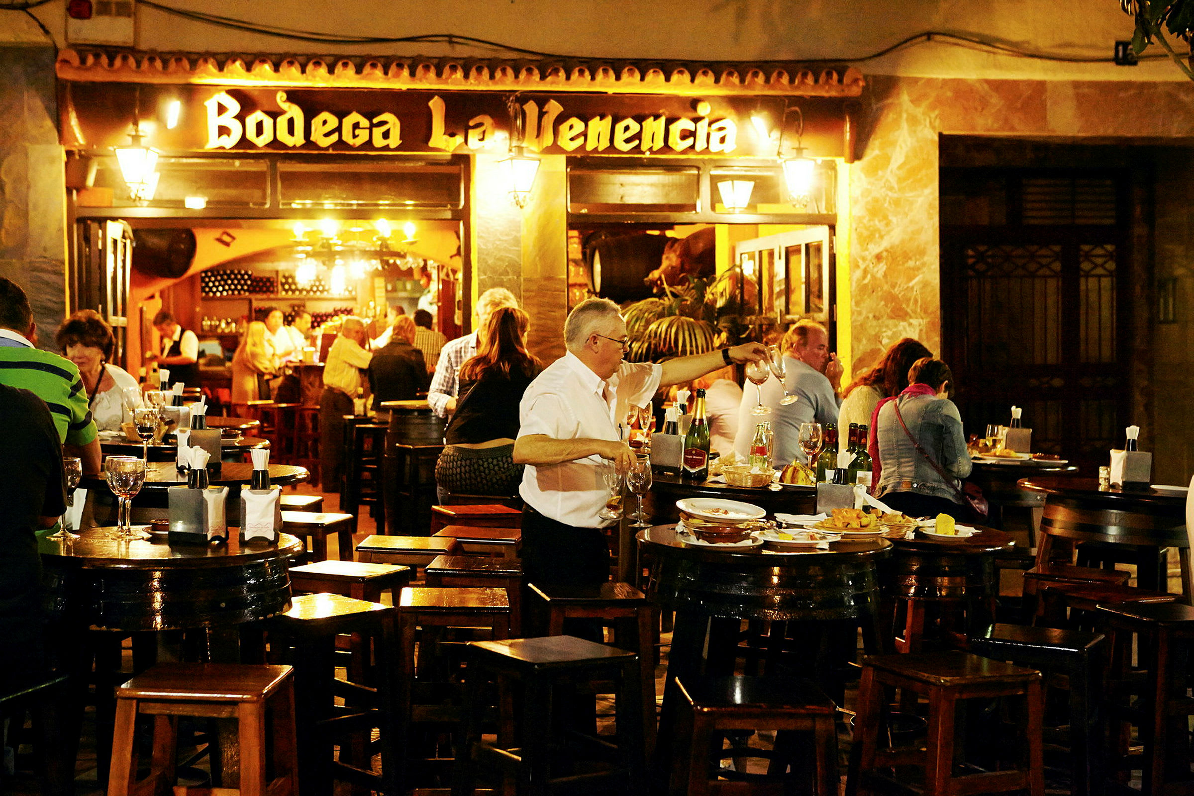 En uteservering med bord og stoler hvor mennesker spiser og drikker. Et skilt med teksten 'Bodega La Venencia' er synlig i bakgrunnen. (AI-generert tekst)