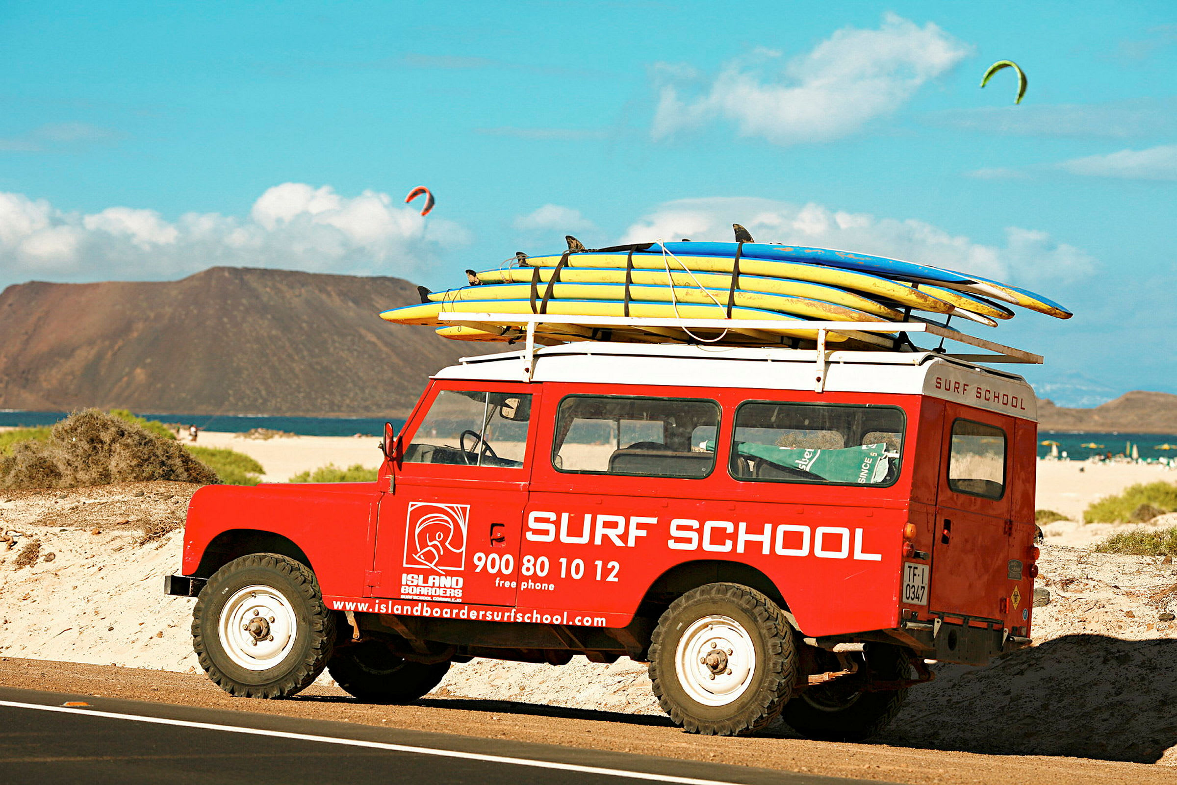 En rød bil merket med "Surf School" står parkert på en strand med surfebrett festet på taket. (AI-generert tekst)