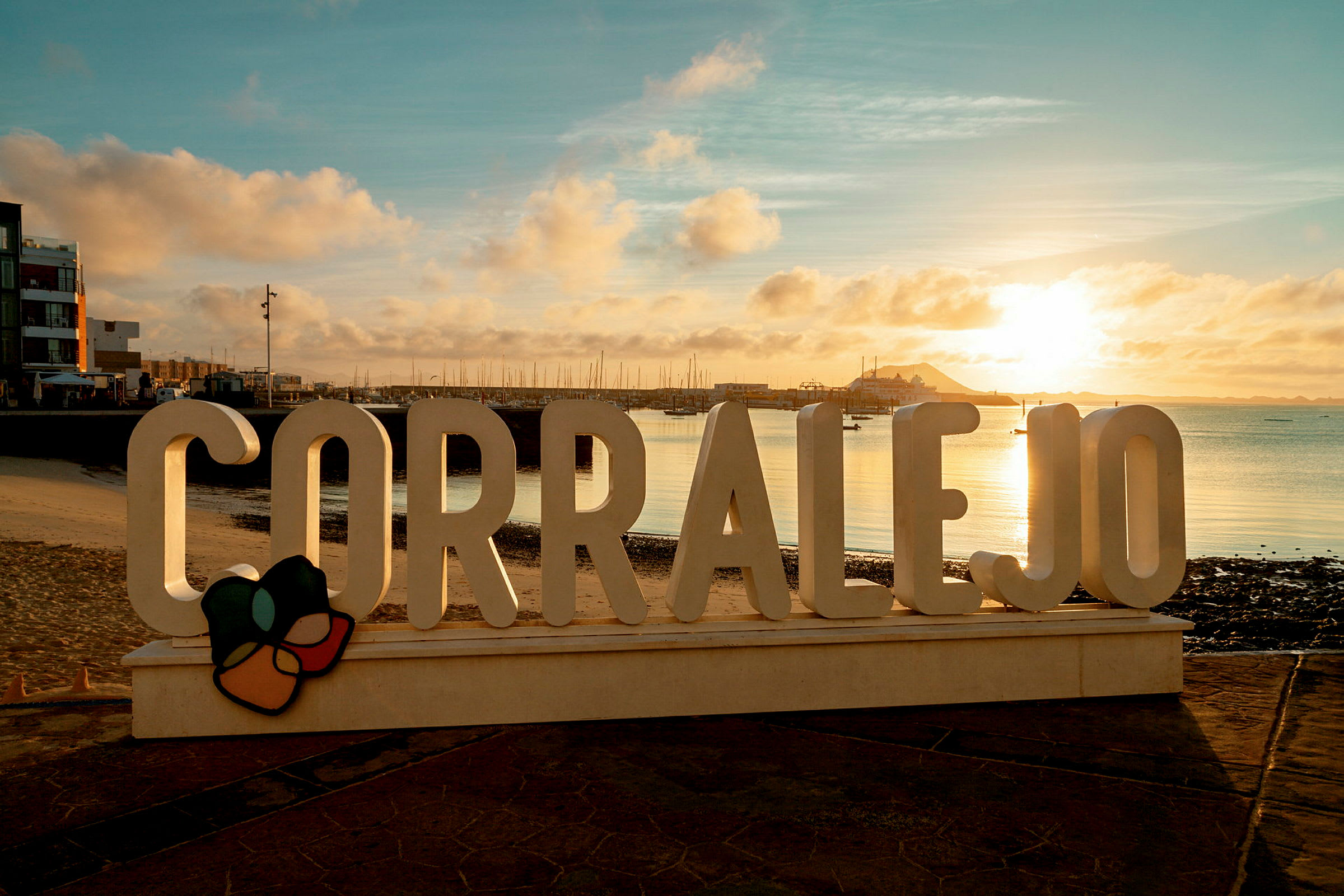 Skiltet 'Corralejo' ved en promenadevei med havet og en kai i bakgrunnen under en solnedgang. (AI-generert tekst)