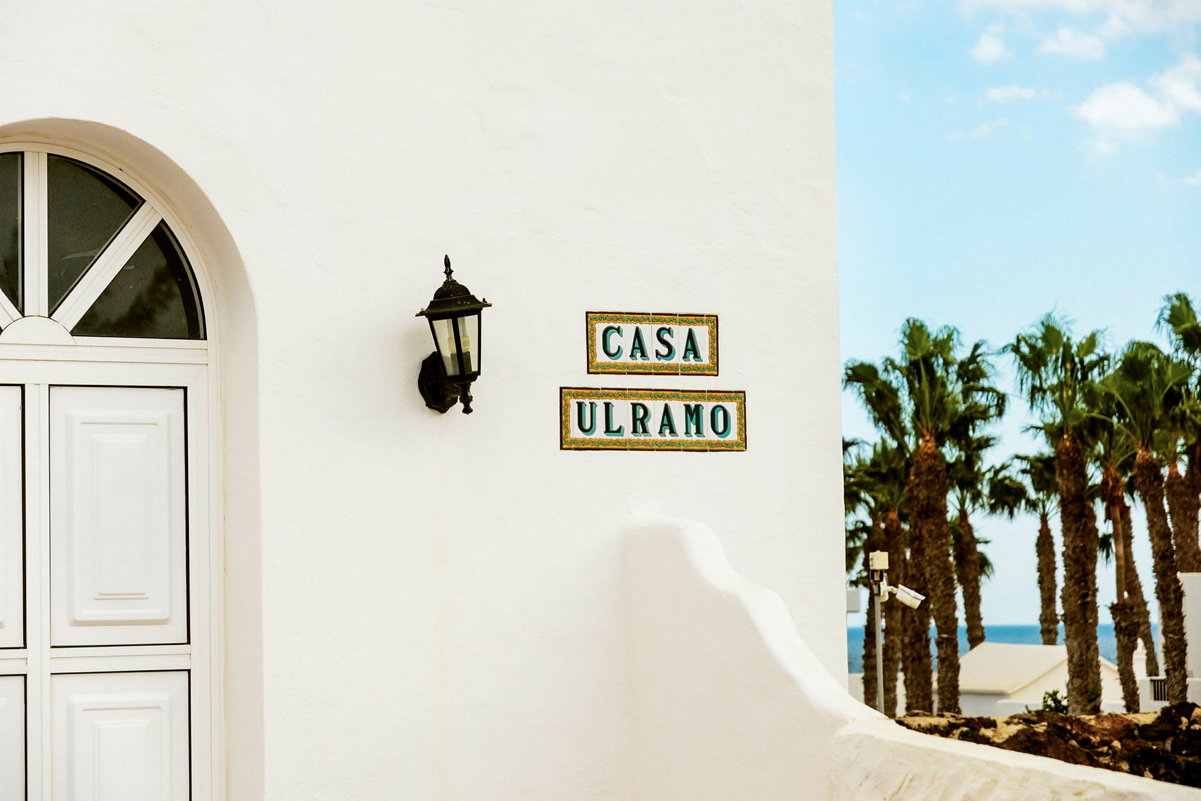 Nærbilde av en hvit bygningvegg med et bueformet vindu, en svart utendørslykt og fliser med teksten 'CASA ULRAMO'. Utsikt over palmer og havet ses i bakgrunnen. (AI-generert tekst)