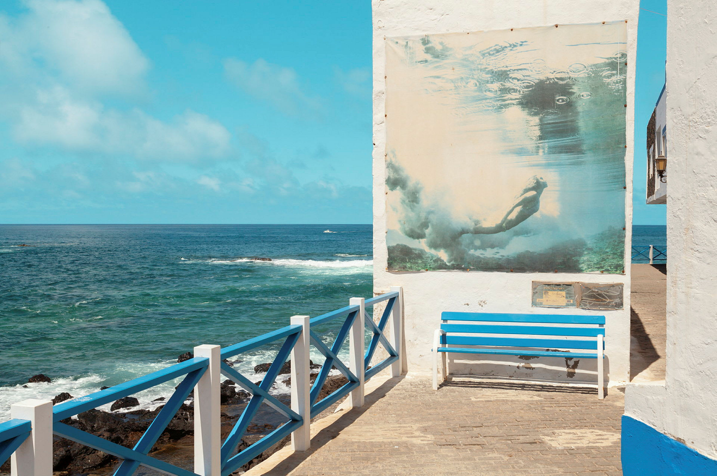 En blå benk ved siden av et hvitmalt rekkverk med utsikt over havet og en vegg med en plakat av en dykker. (AI-generert tekst)