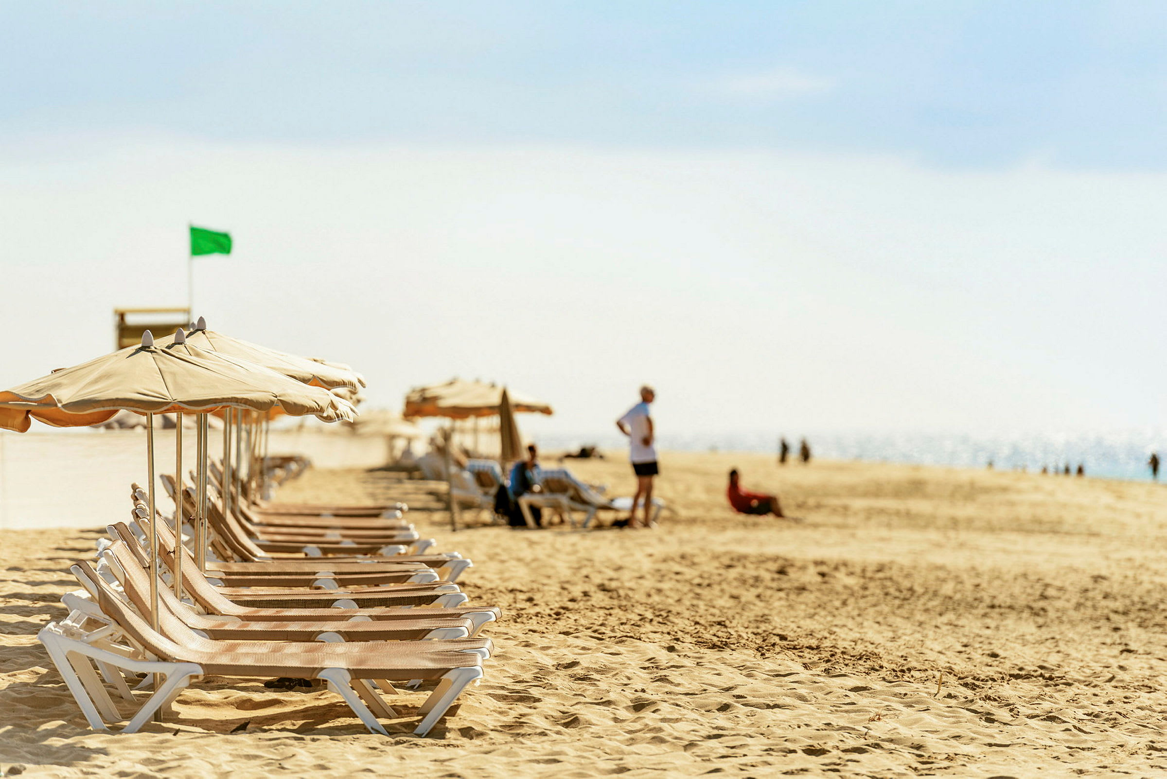 Solstoler med parasoller på en sandstrand, noen mennesker ses i bakgrunnen. (AI-generert tekst)