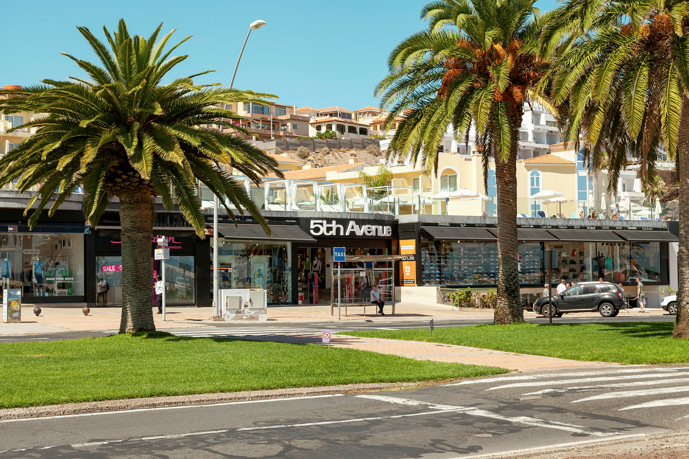 Et shoppingområde med palmer, grønt gress og en butikk med skiltet "5th Avenue". (AI-generert tekst)