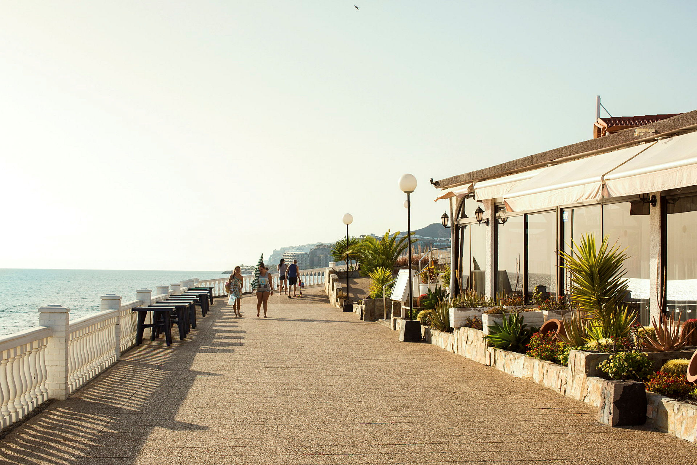 Strandpromenade med havutsikt, bord langs rekkverket og folk som går tur. (AI-generert tekst)