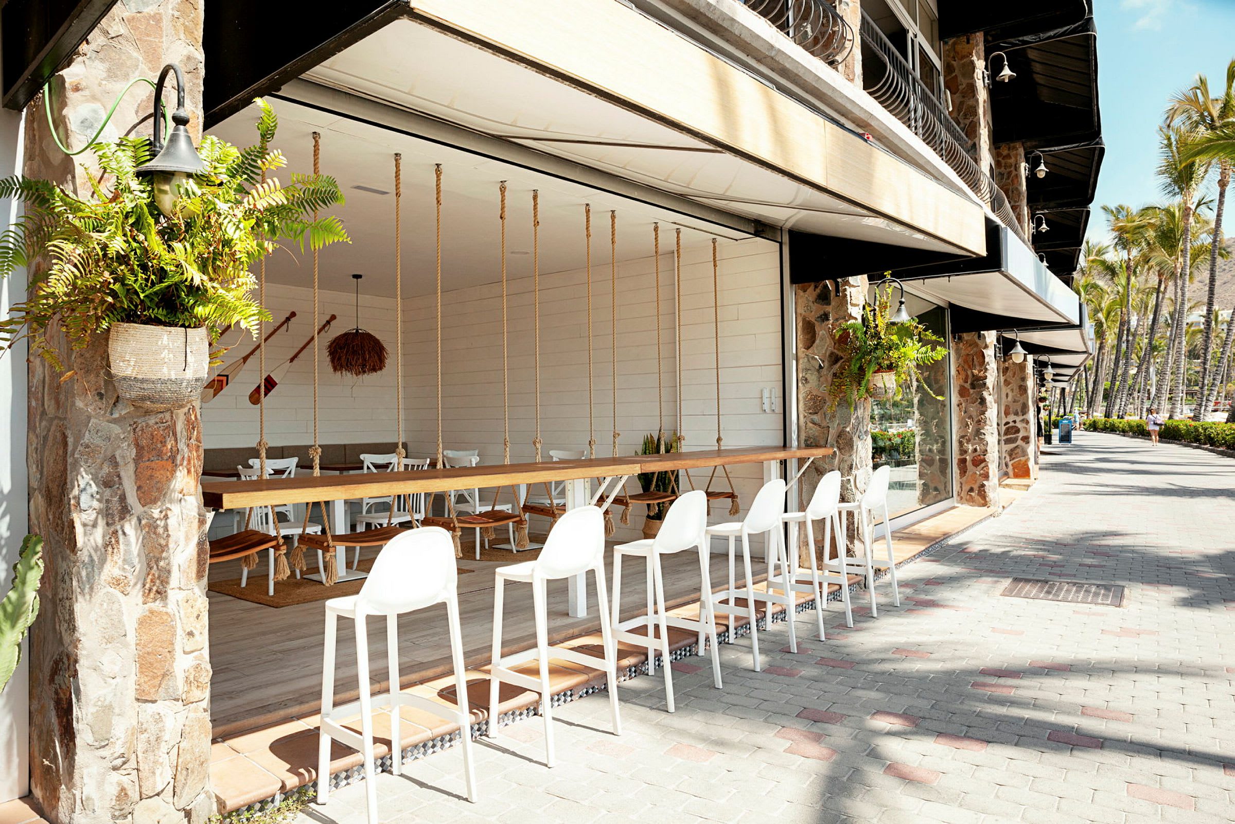 Utendørsområde med hvite barstoler ved en benk, med plantepynt og en restaurant med sitteplasser i umiddelbar nærhet. (AI-generert tekst)
