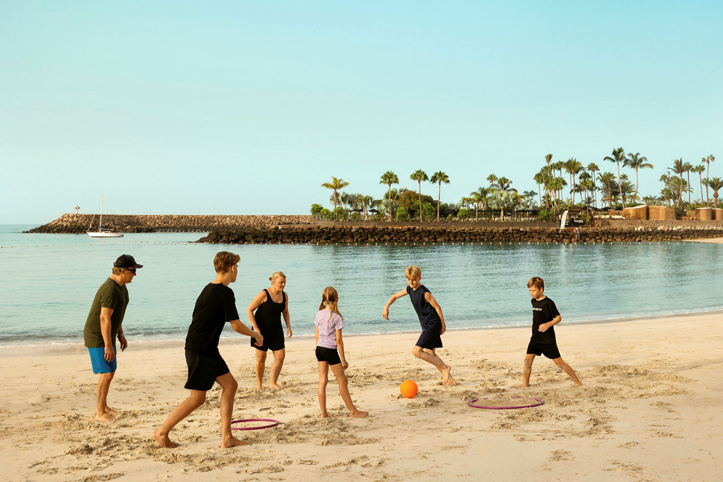 En gruppe personer leker med en ball på en sandstrand, med en havn og palmer i bakgrunnen. (AI-generert tekst)