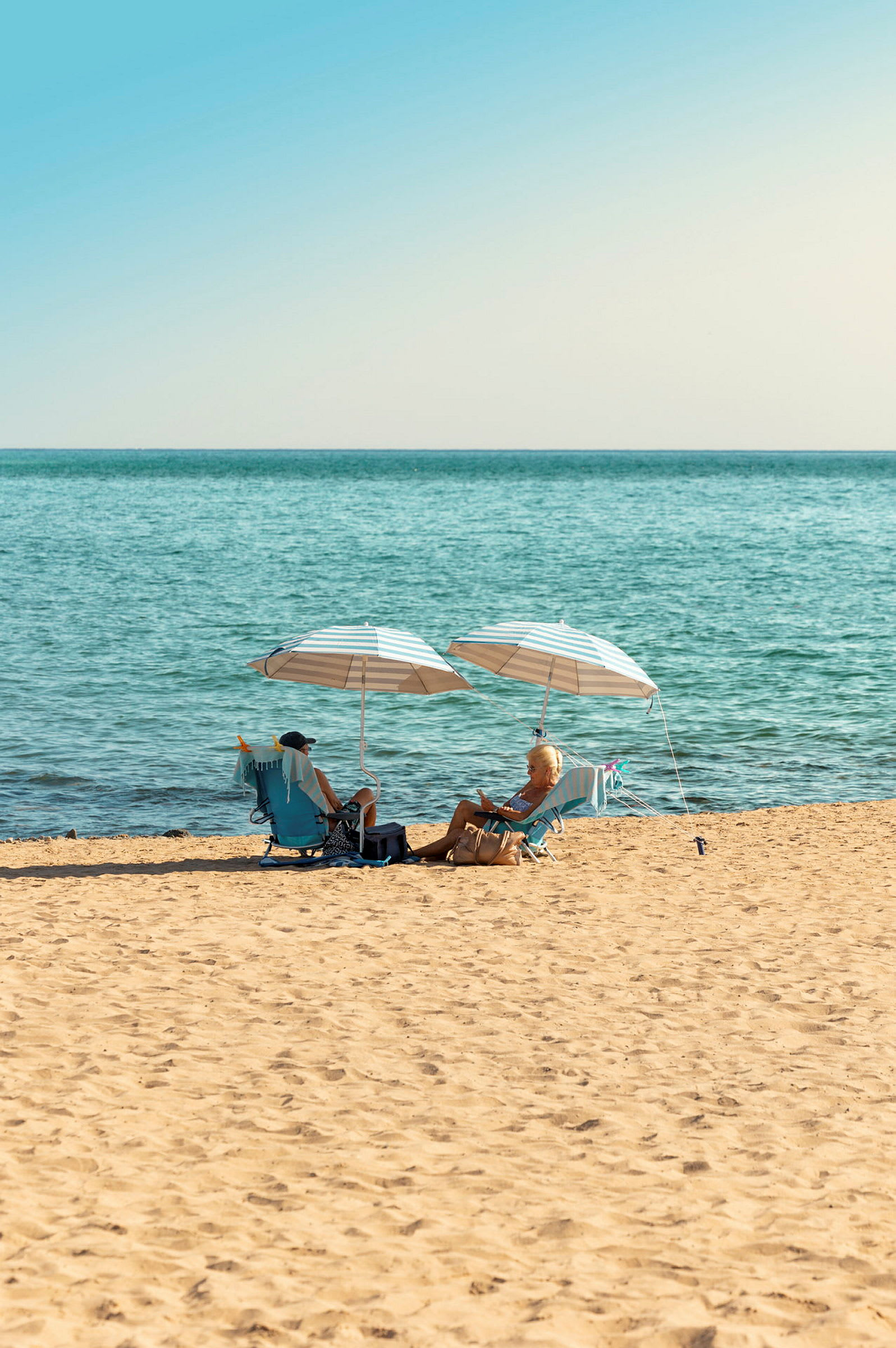En strand med to personer som sitter under parasoller og nyter utsikten over havet. (AI-generert tekst)
