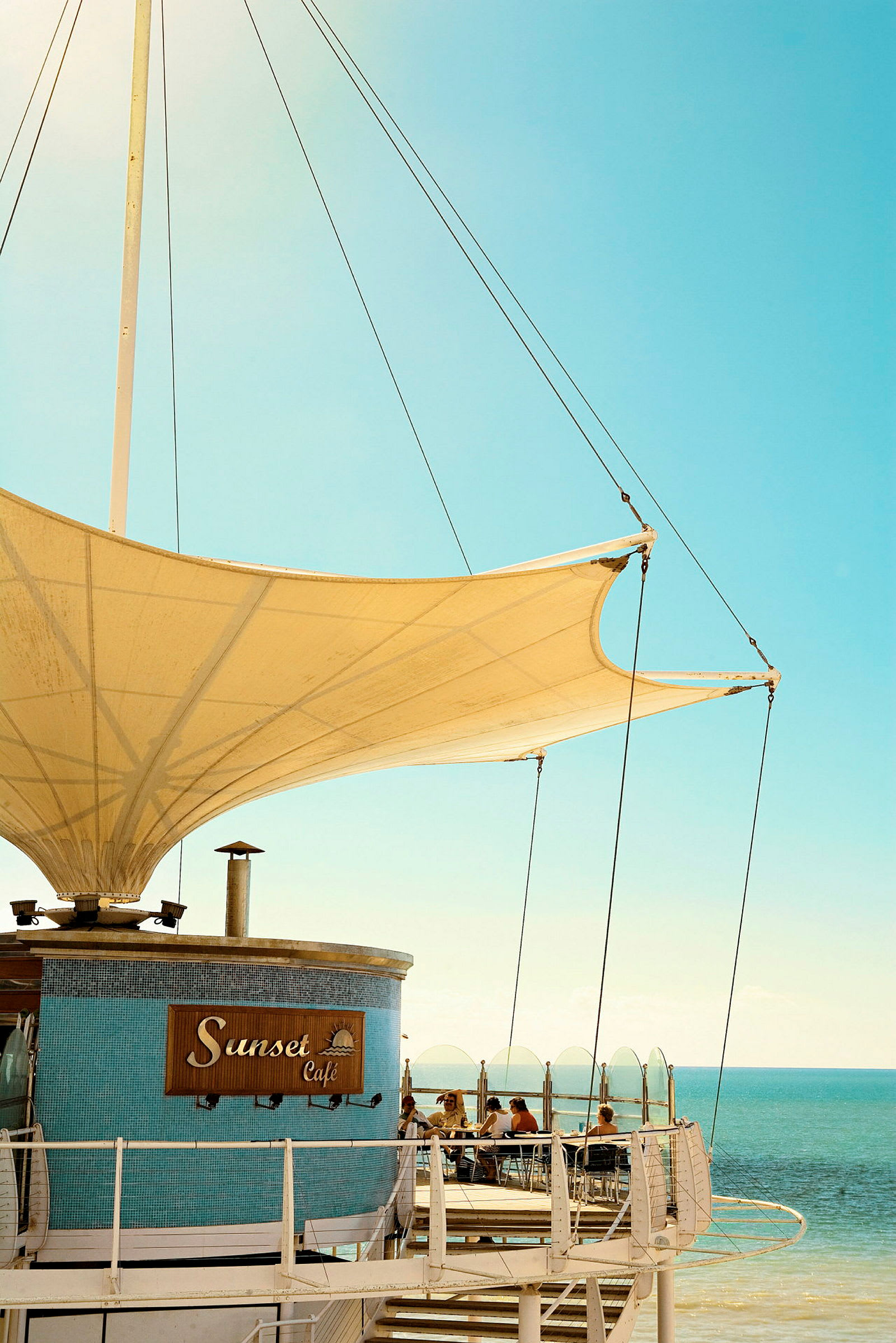 Bilde av Sunset Café med havet i bakgrunnen, sittende gjester og et stort seillignende tak. (AI-generert tekst)
